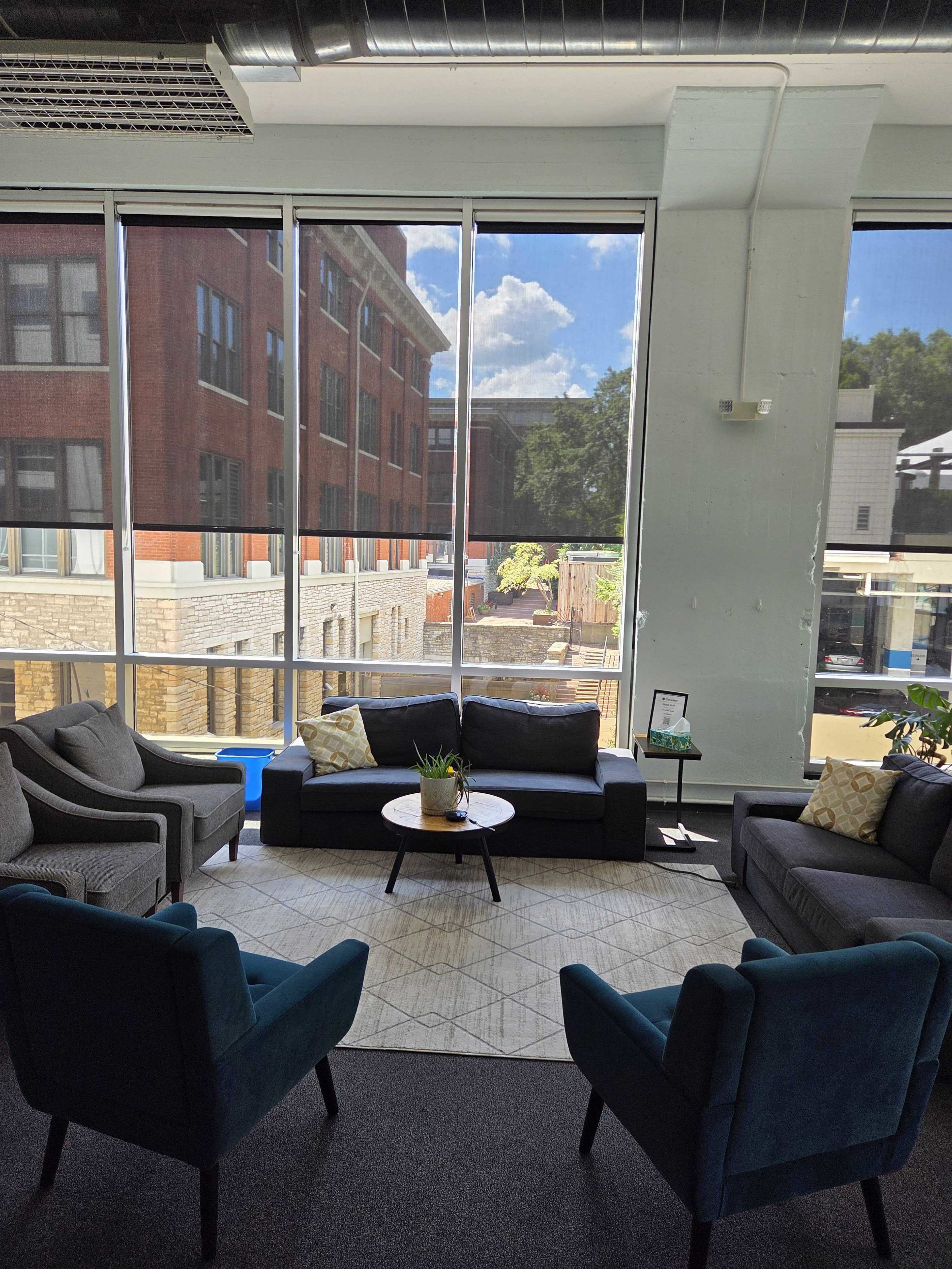 The image shows a modern lounge area with gray sofas, teal chairs, and large windows allowing natural light to illuminate the space.