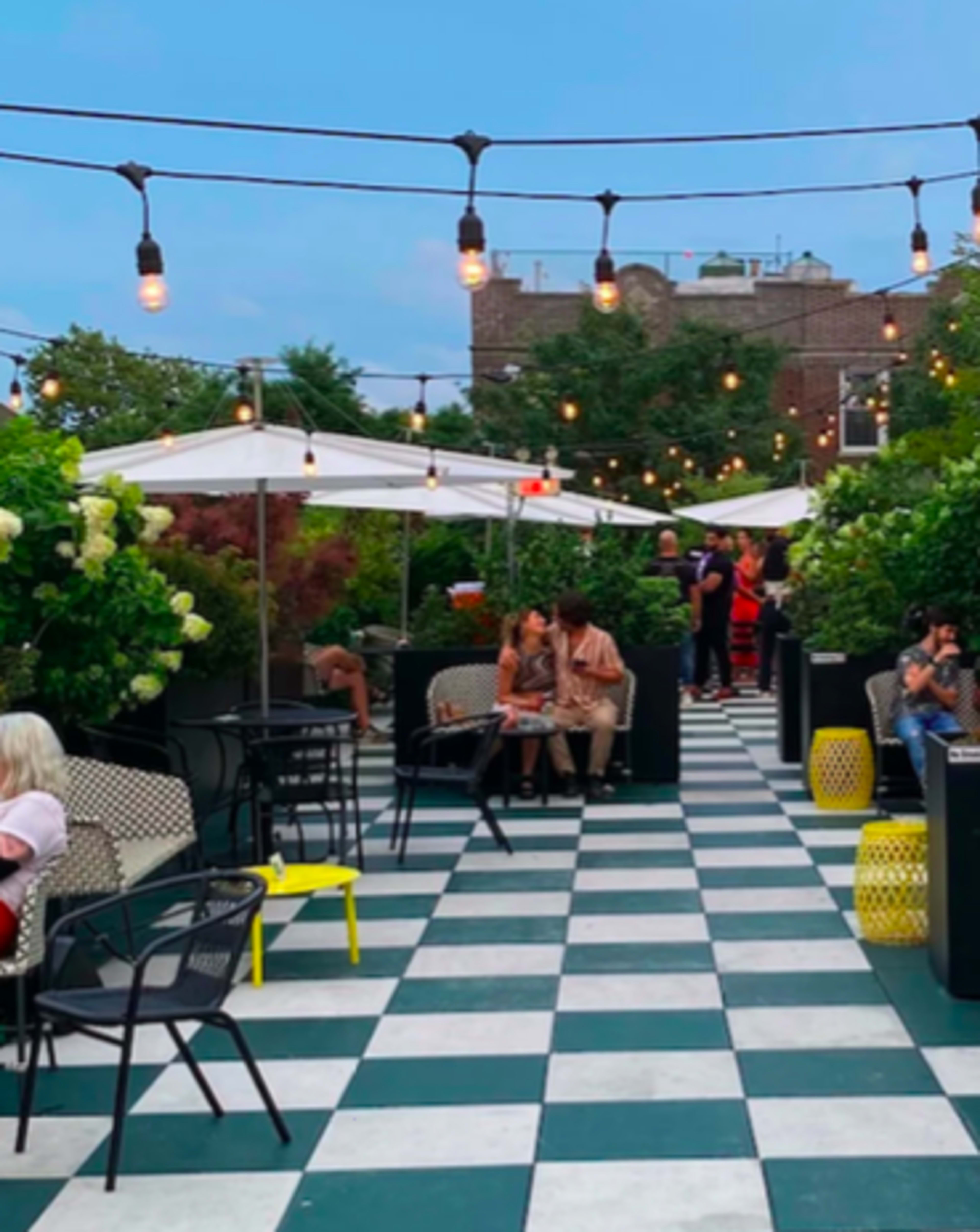 The image shows a rooftop lounge area with checkered flooring, string lights overhead, and groups of people seated at tables among greenery and umbrellas.