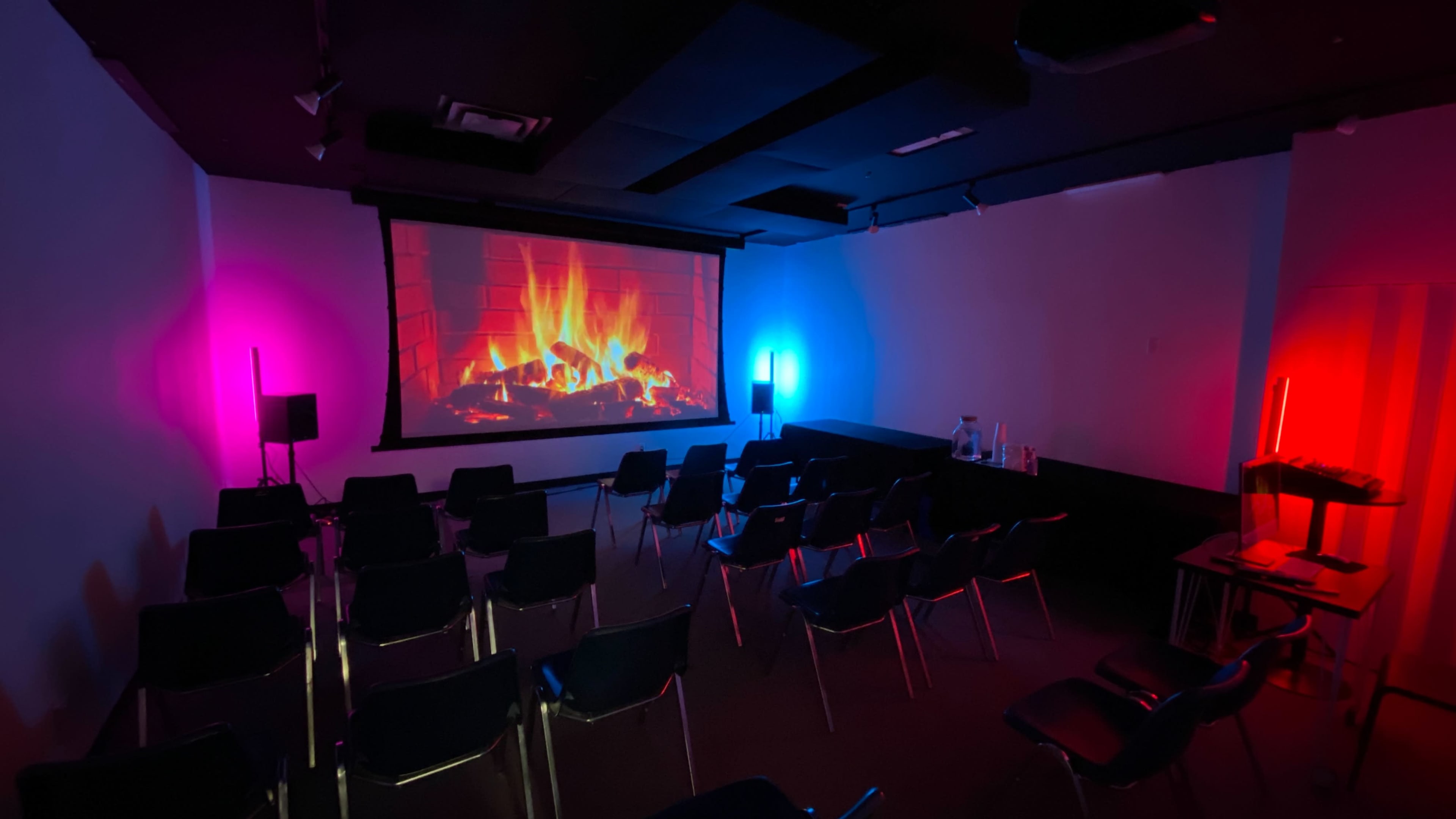 A room with black chairs arranged in rows faces a large screen displaying a digital fireplace, illuminated by colorful lights on the walls.