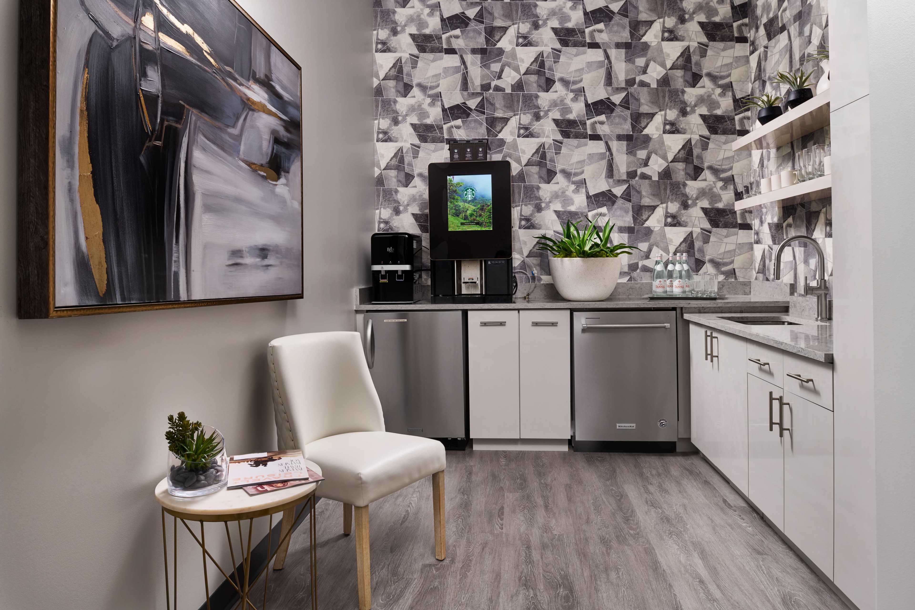 A modern kitchenette featuring a water dispenser, a refrigerator, a sink, and minimalist cabinetry against a geometric patterned wall, with a chair and a small table placed nearby.
