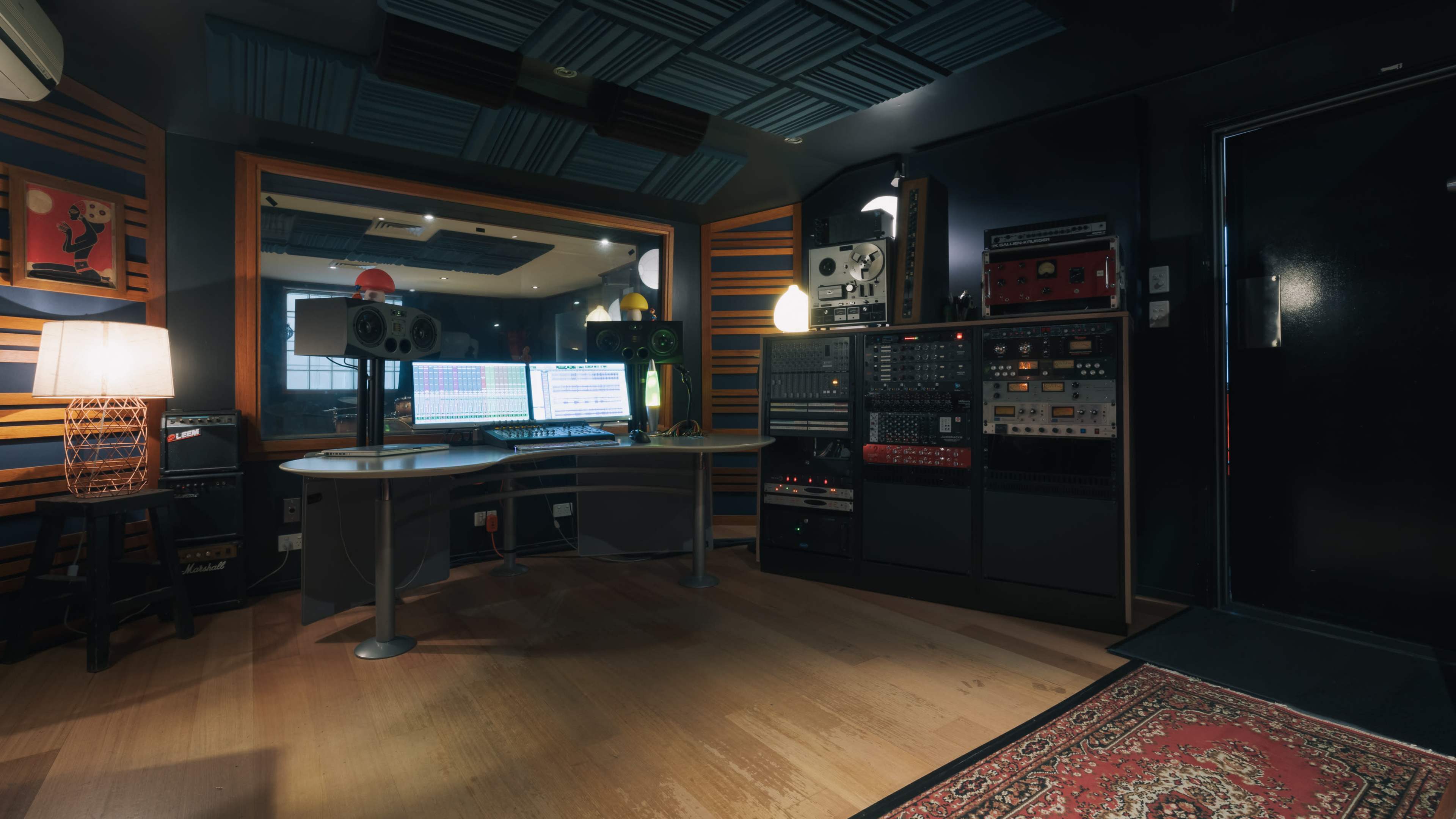 A music recording studio features a mixing desk surrounded by audio equipment, speakers, and a decorative lamp.