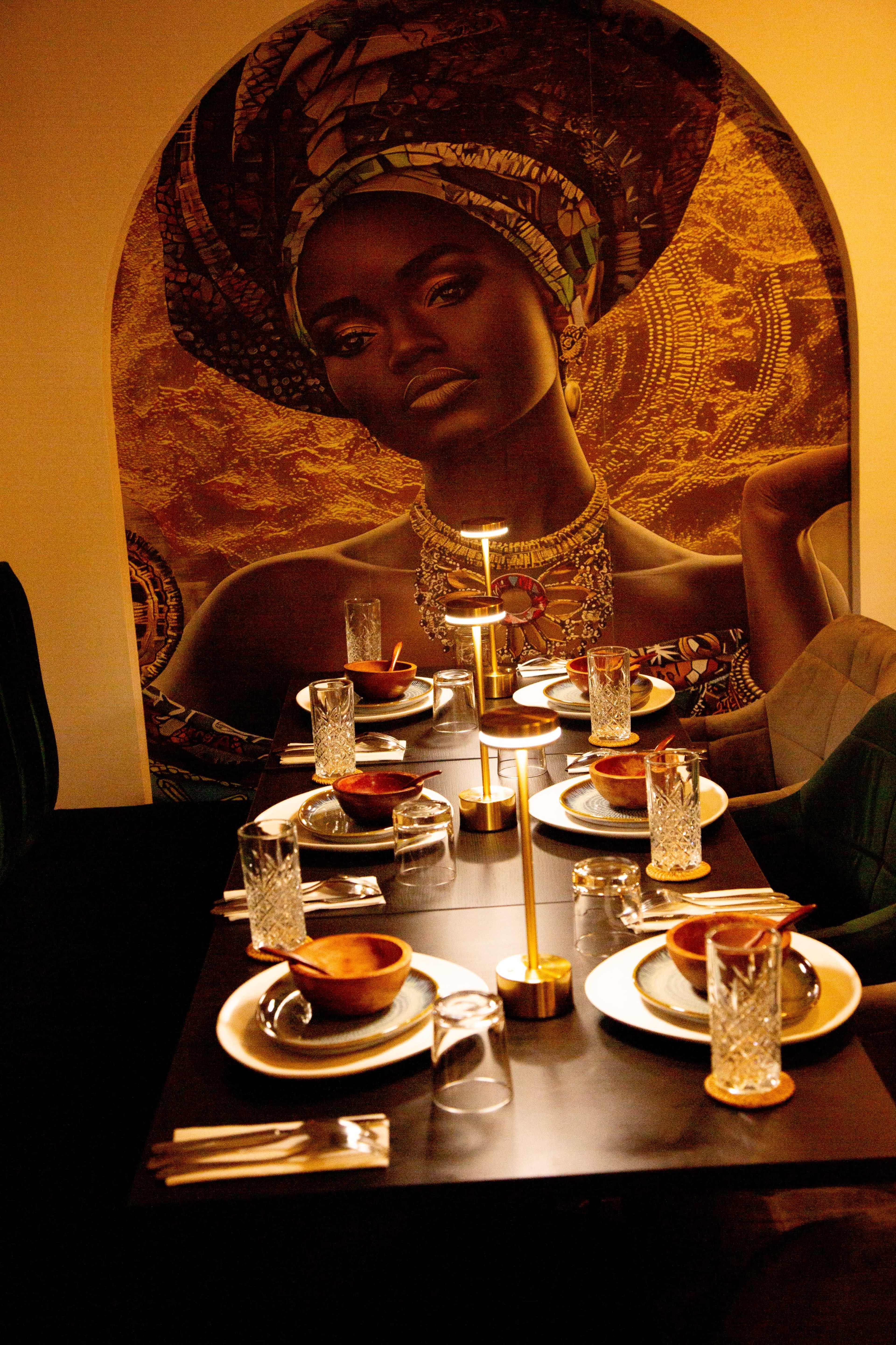 A dining table is elegantly set with plates, cutlery, and glasses, in front of a striking mural depicting a woman's portrait.