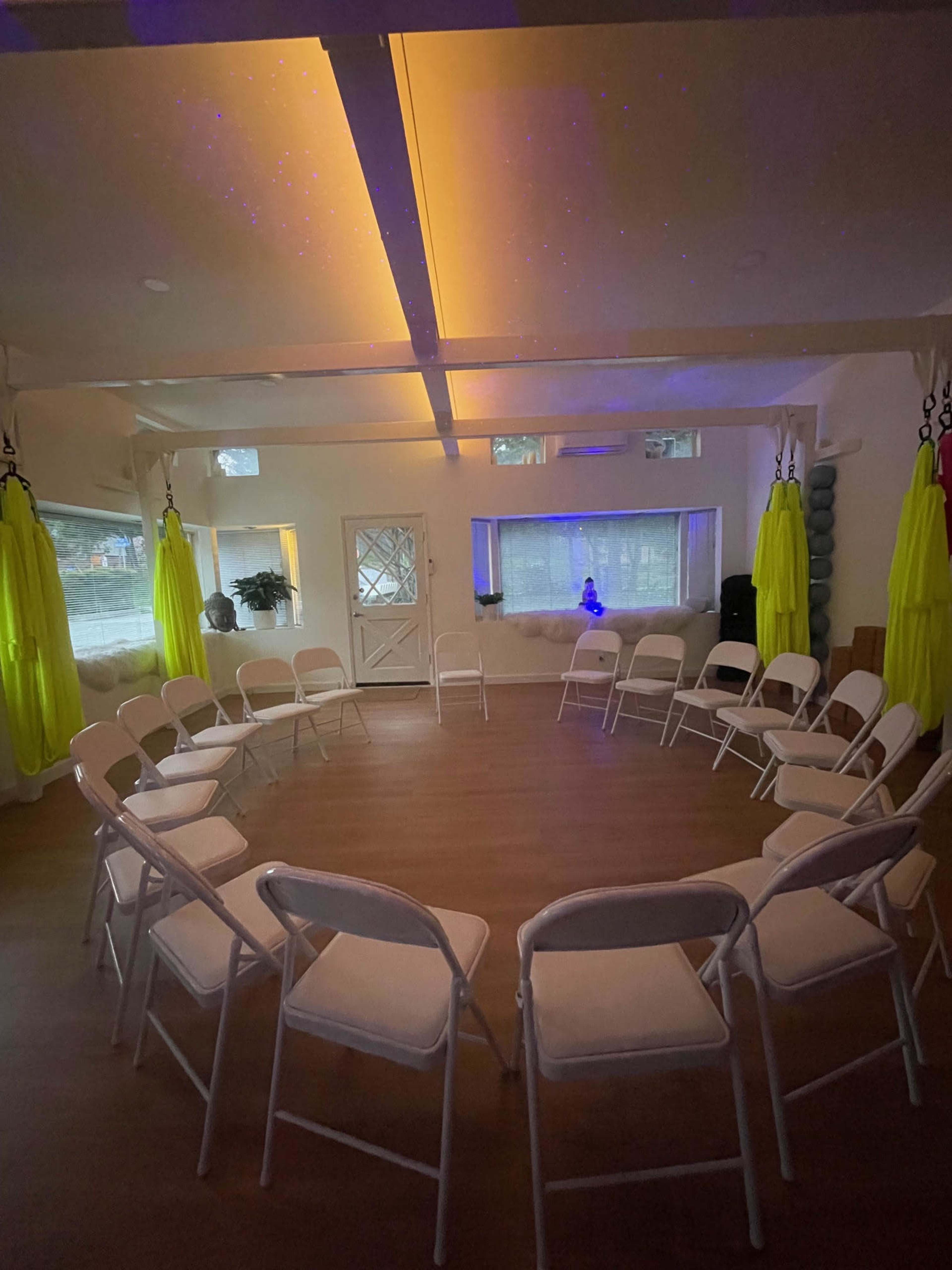 The image shows a circular arrangement of white folding chairs set up in a bright room with green drapes and ambient lighting.