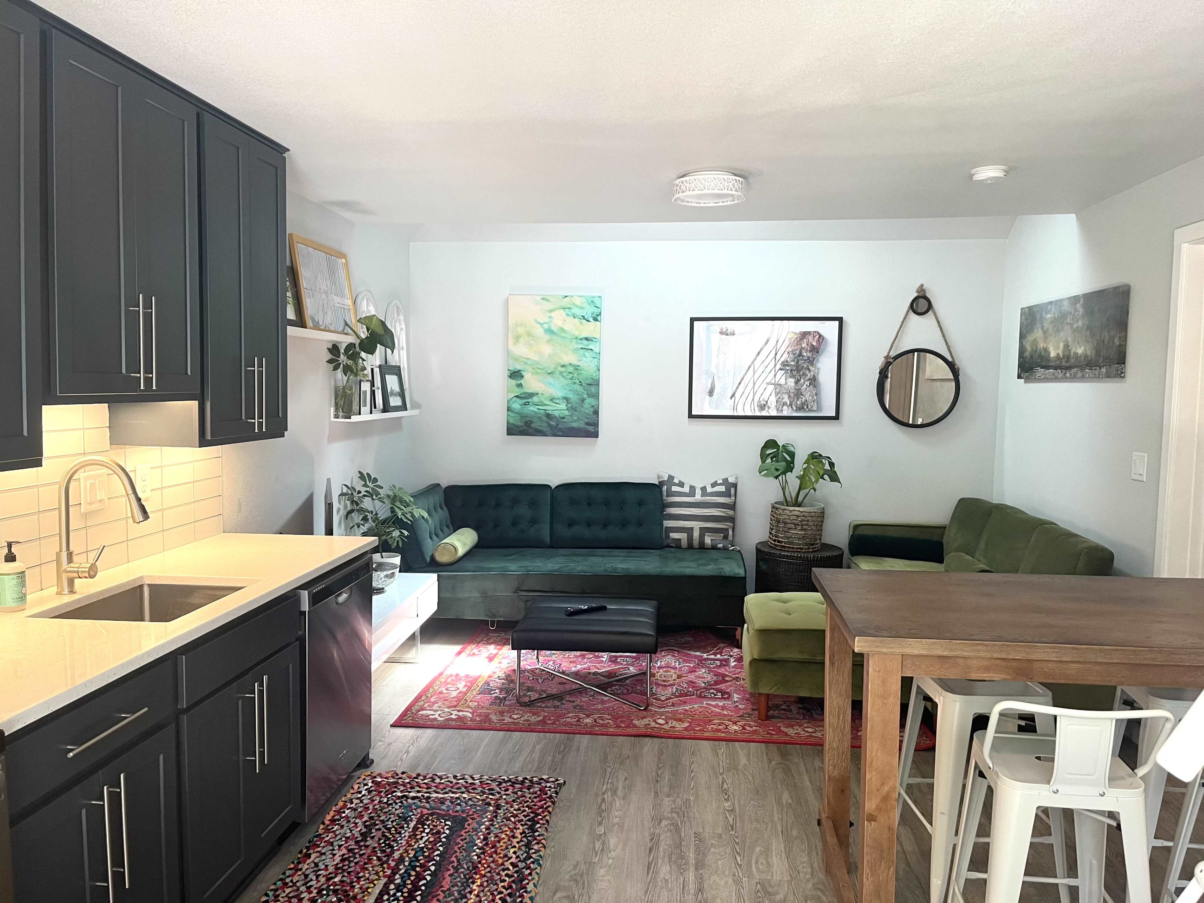 A modern living area features a green sectional sofa, a dining table, and a kitchen with dark cabinetry and a white backsplash.