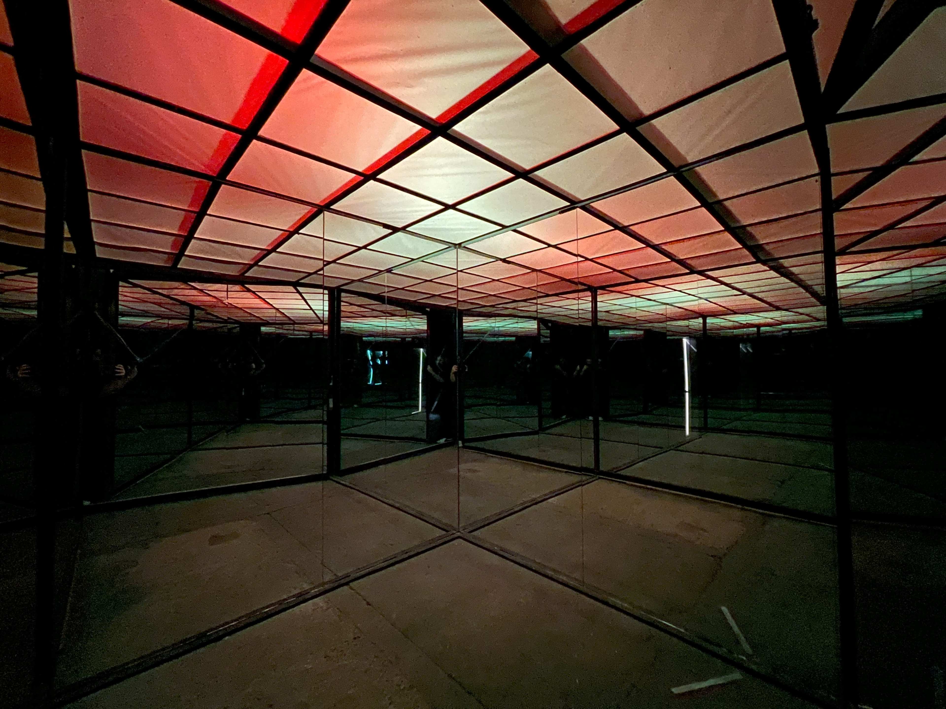 A mirror maze with a grid-patterned ceiling illuminated by red and green lights.