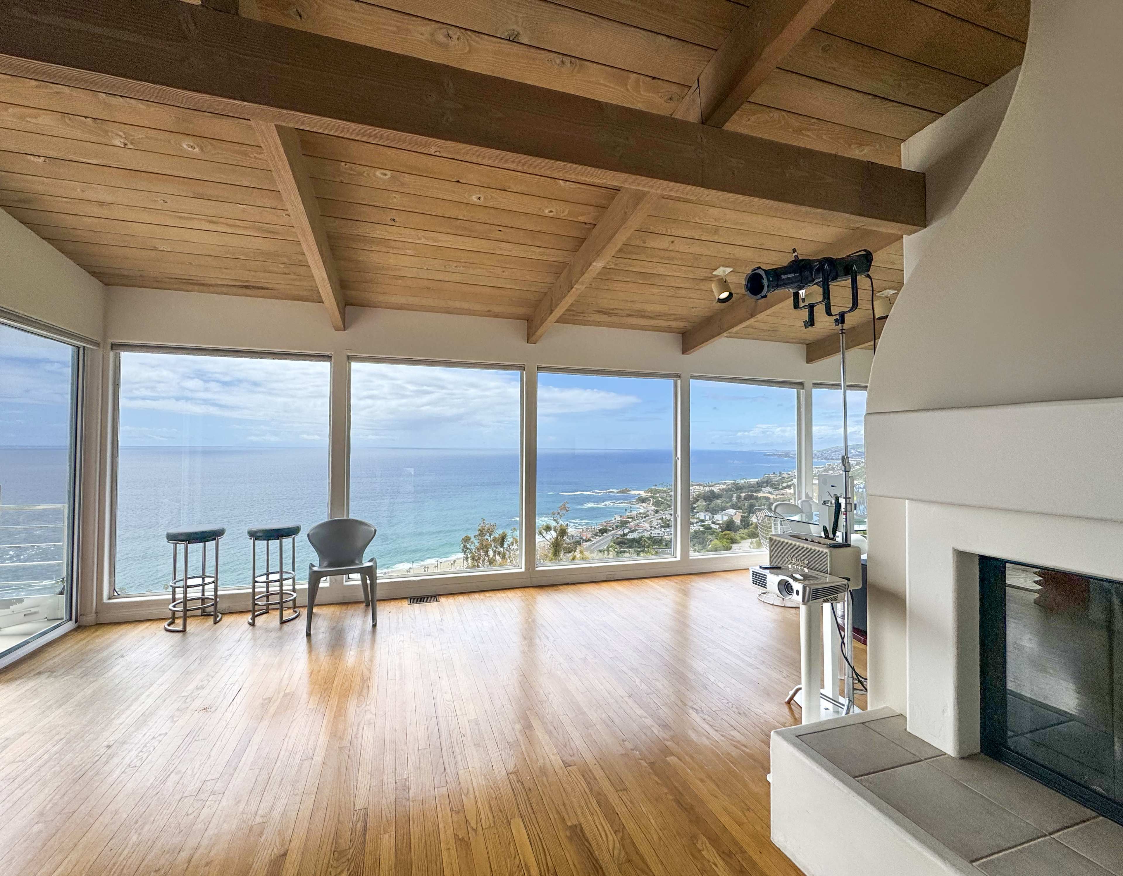 A spacious, empty living room features large windows overlooking the ocean, with wooden beams on the ceiling and a fireplace in the corner.