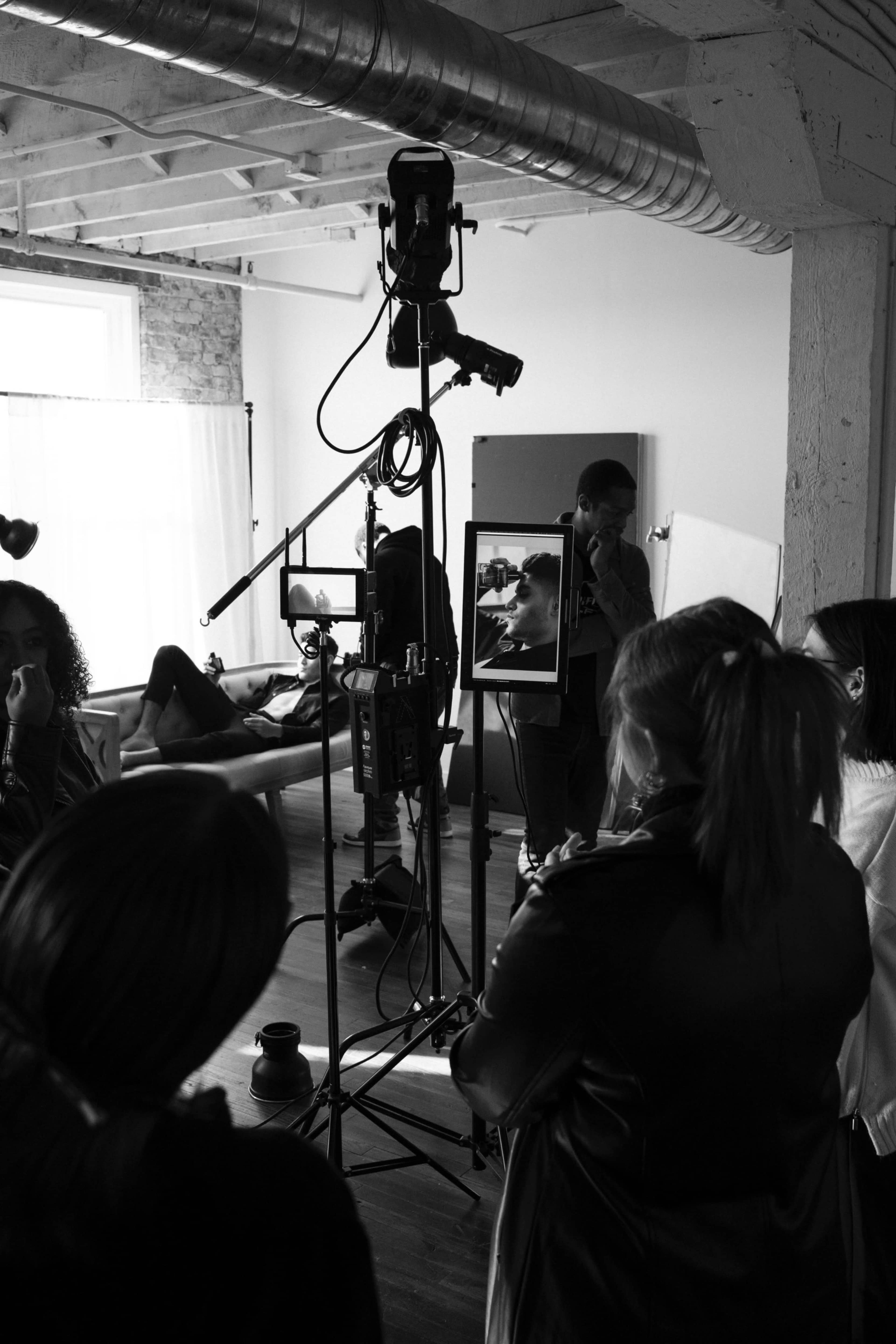 A group of people are gathered around a camera setup in a well-lit studio, while one individual is being styled or prepared for a shoot.
