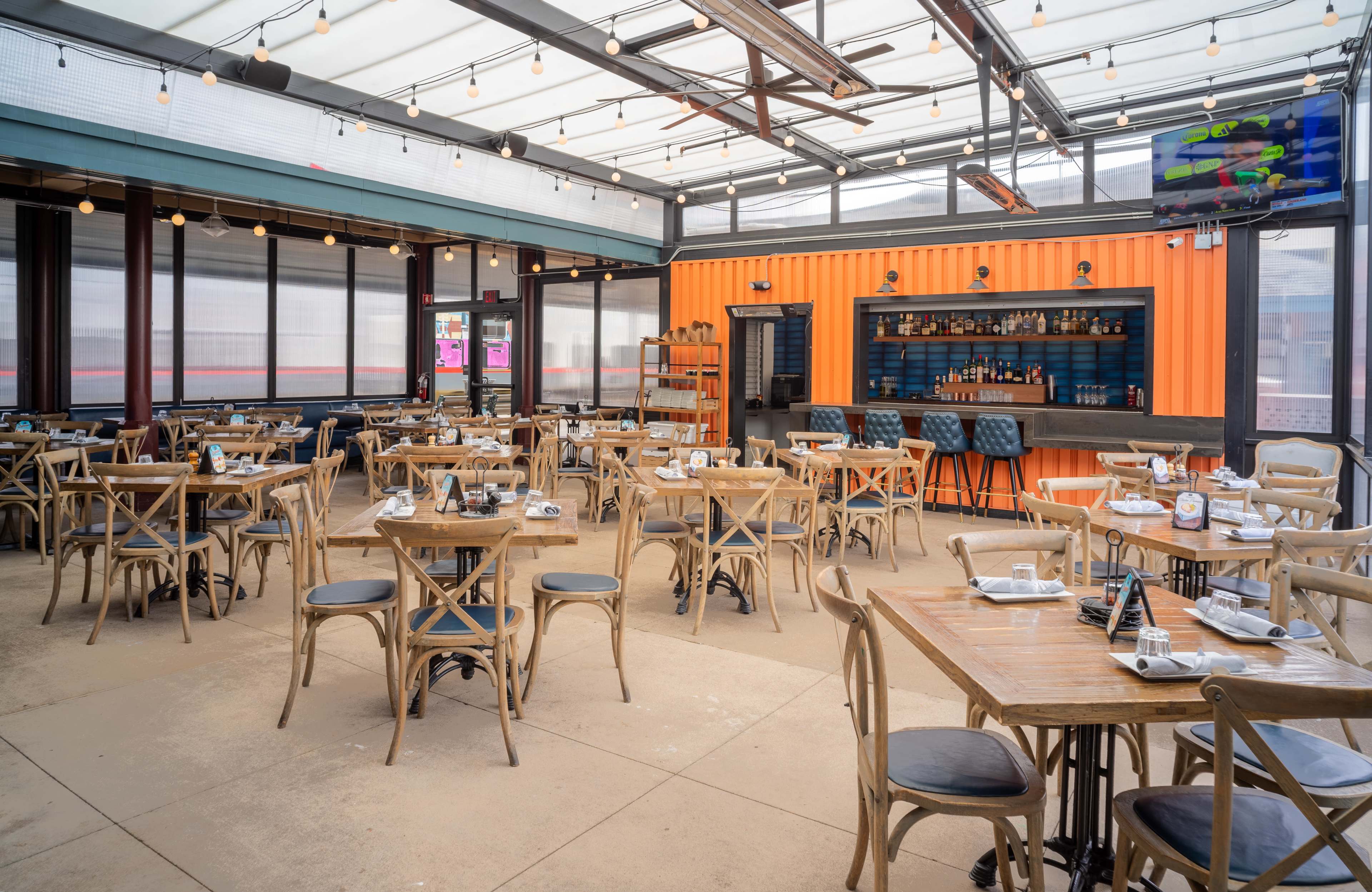 The image shows a spacious restaurant with wooden tables and chairs, featuring a bar area in an orange shipping container and large windows allowing natural light.