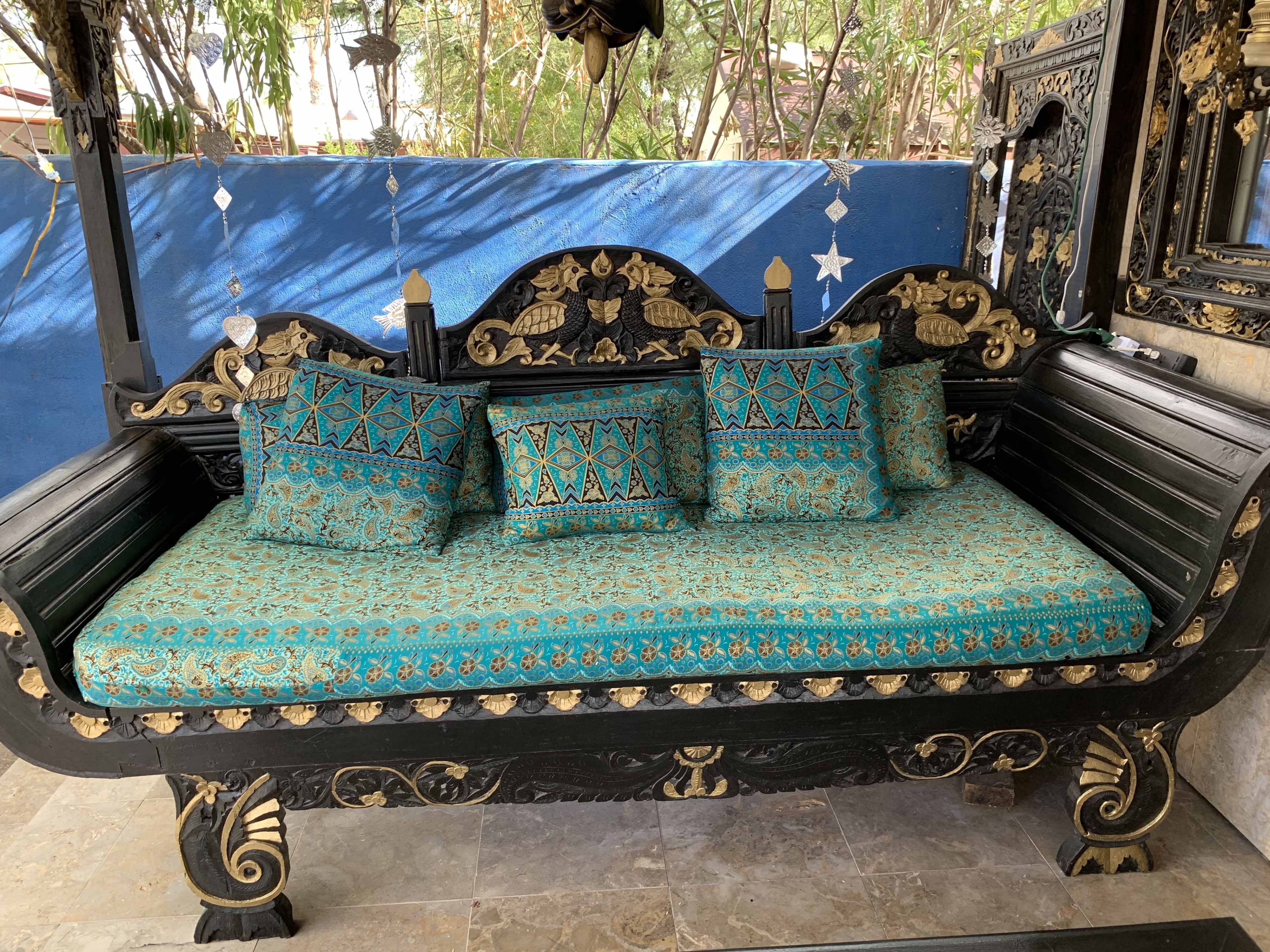 A black wooden bench with intricate carvings, topped with a turquoise upholstered cushion and four matching decorative pillows.