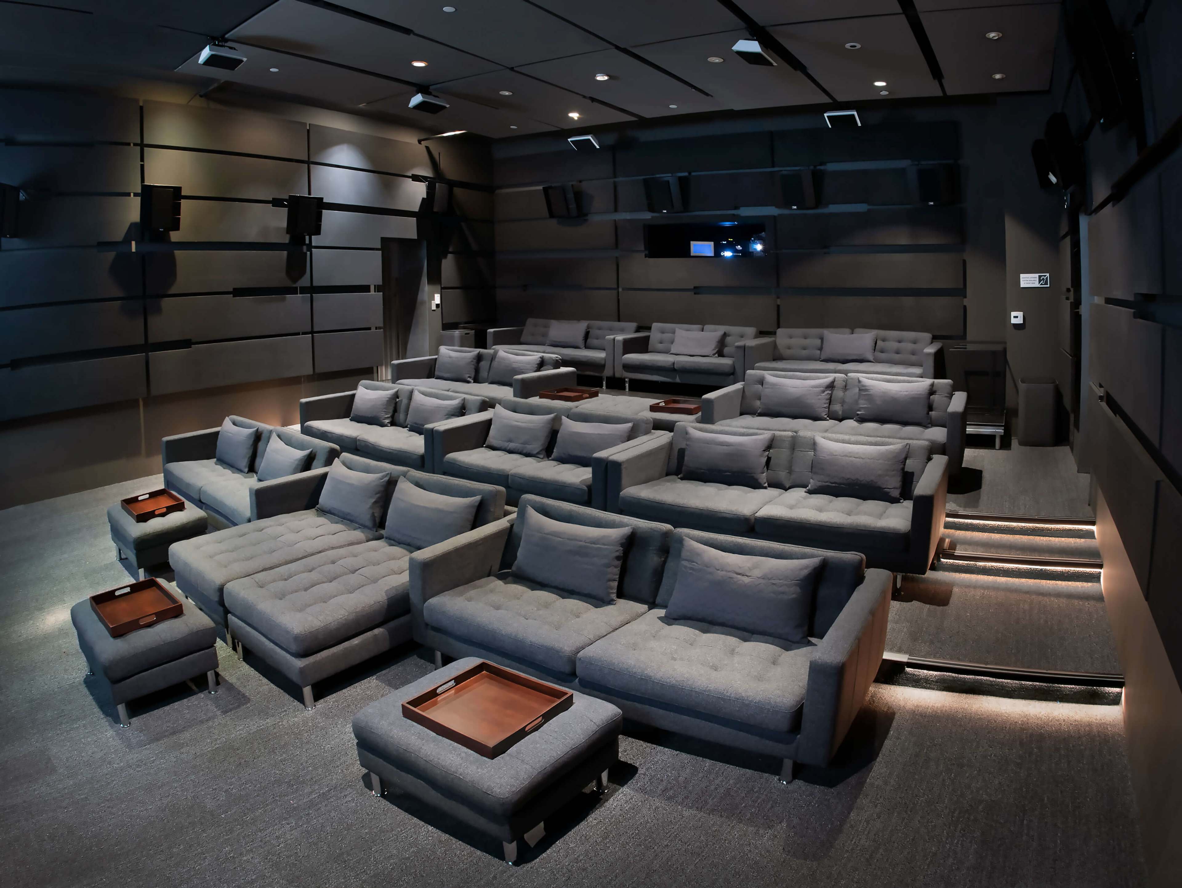 The image shows a modern home theater with multiple gray sectional sofas arranged in tiered levels, facing a wall-mounted screen.