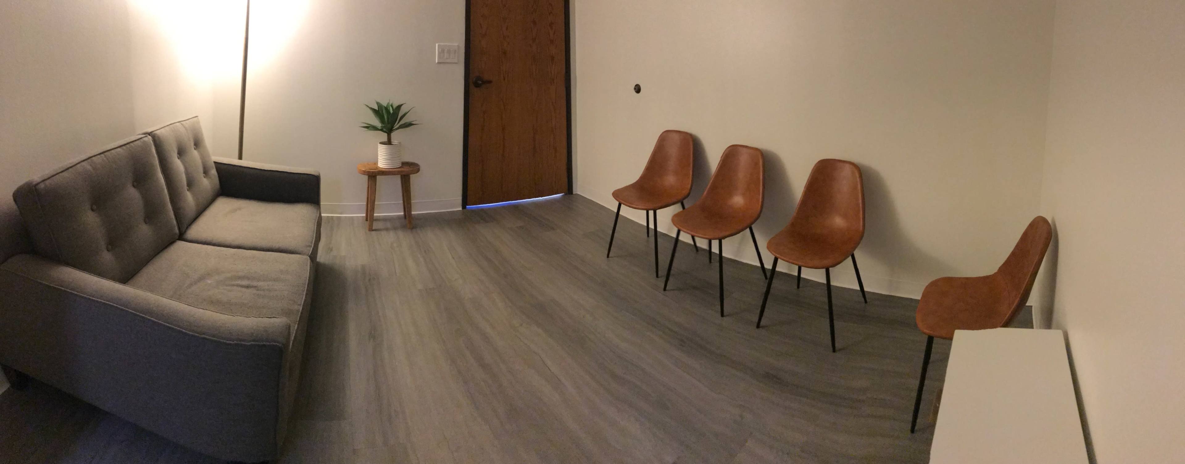 A minimalist waiting room with a gray sofa, four brown chairs, a small table with a plant, and a wooden door.