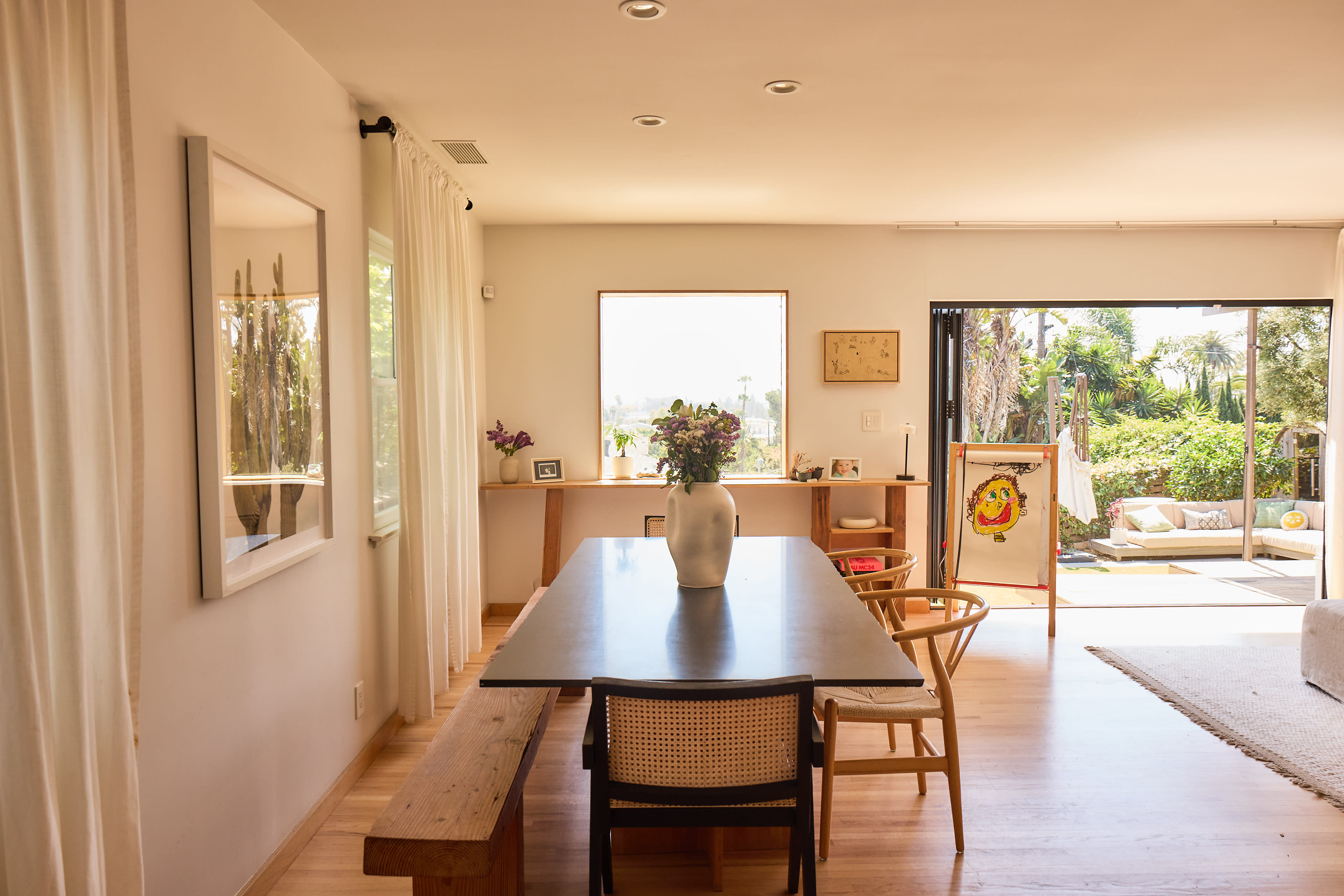A bright dining area features a long table with chairs, a large window overlooking a garden, and light-colored walls.