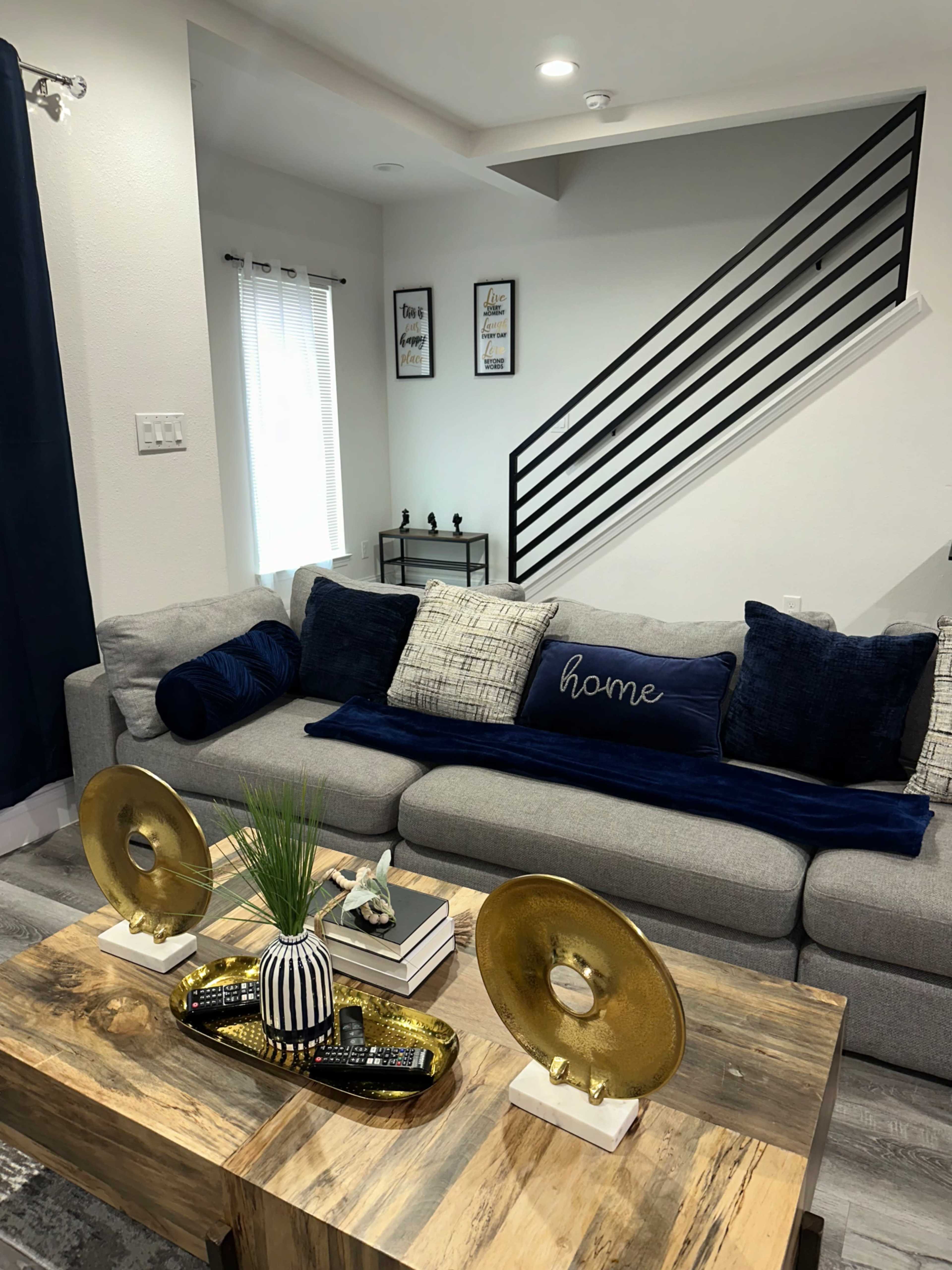 A modern living room featuring a gray sofa adorned with decorative pillows, a wooden coffee table with a decorative tray, and a staircase in the background.