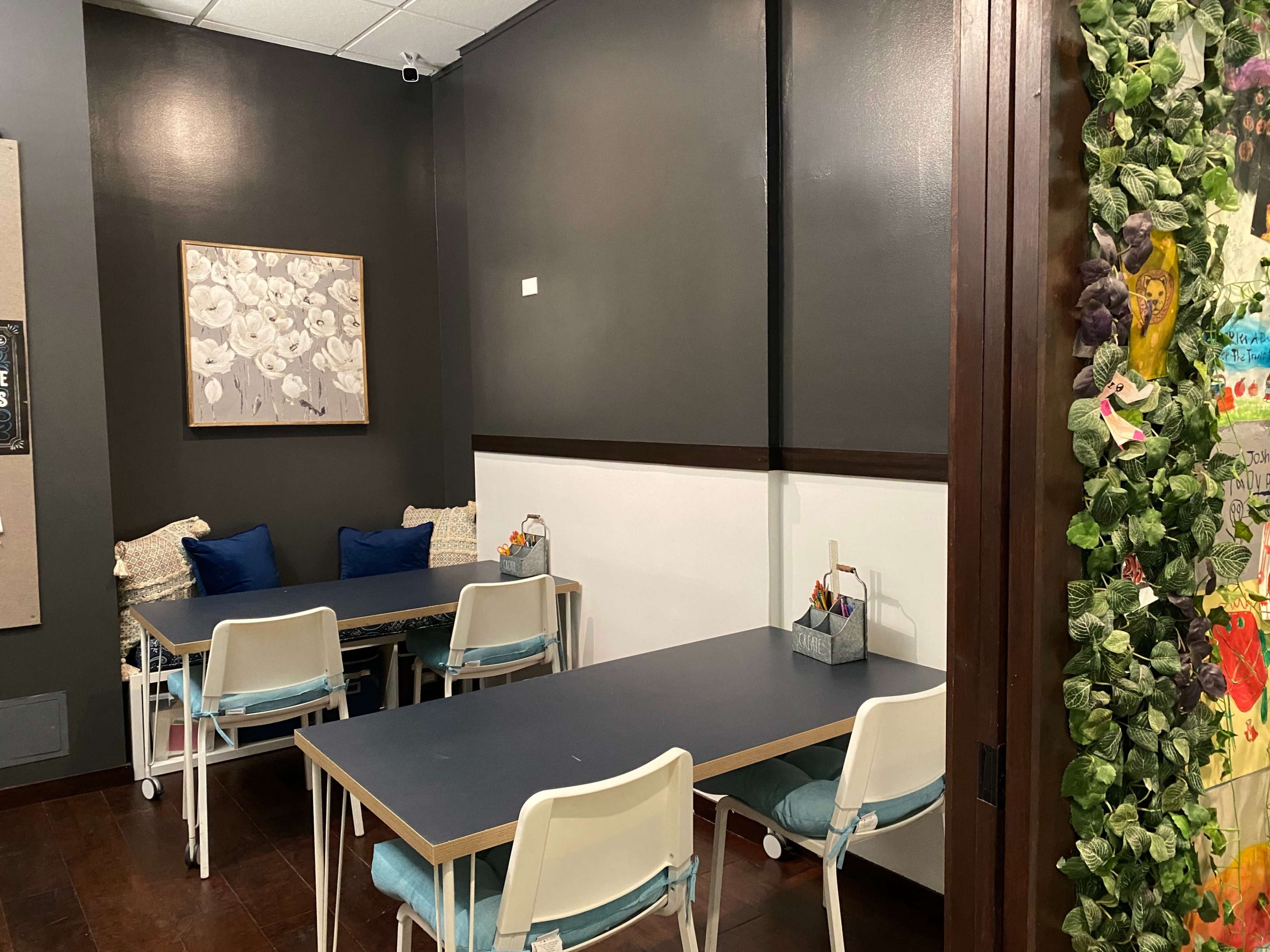 A small meeting room features two tables with white chairs and blue cushions, alongside a wall adorned with decorative plants and framed artwork.