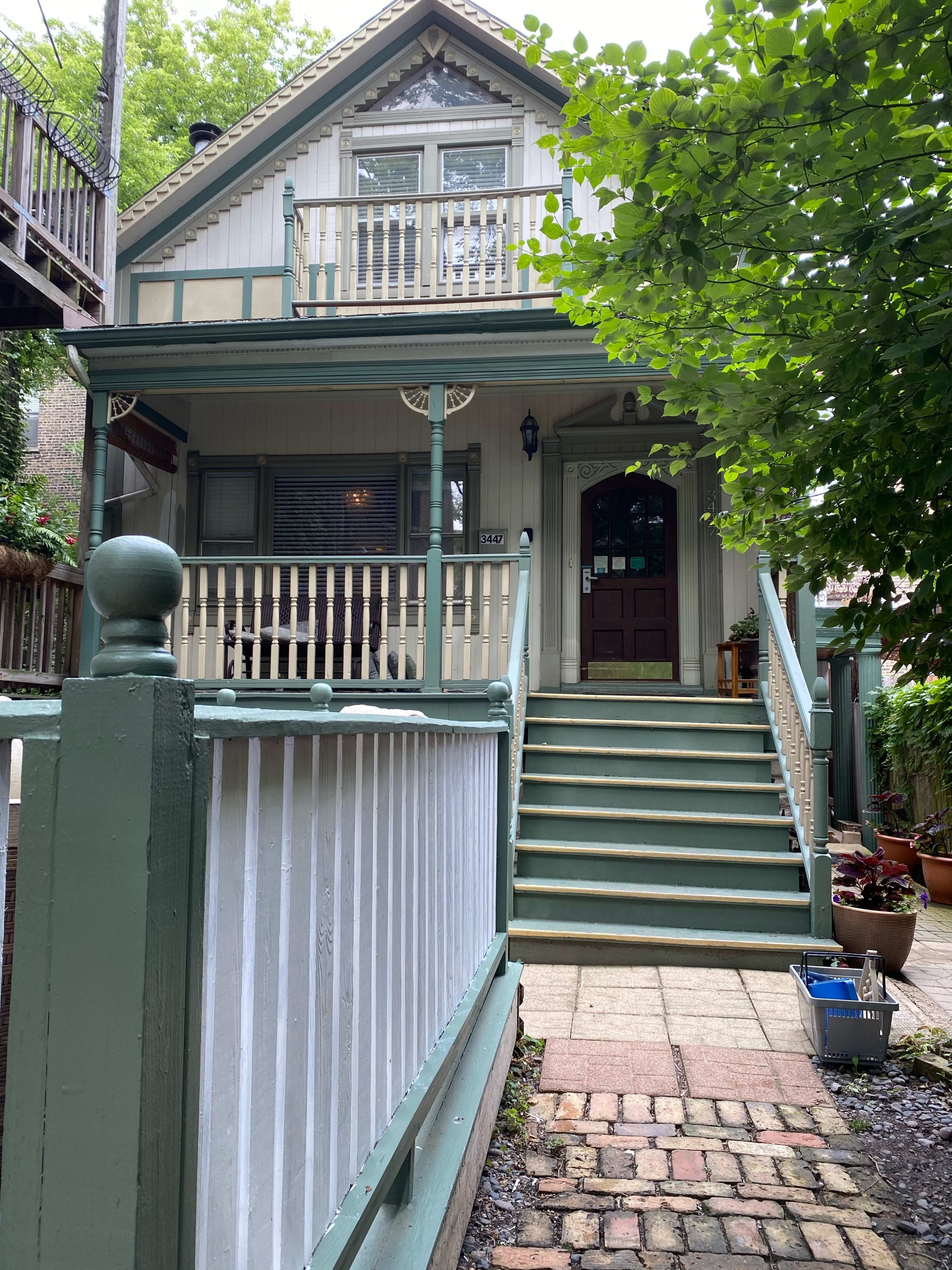 A two-story house with a front staircase and a porch, framed by greenery and a brick pathway.