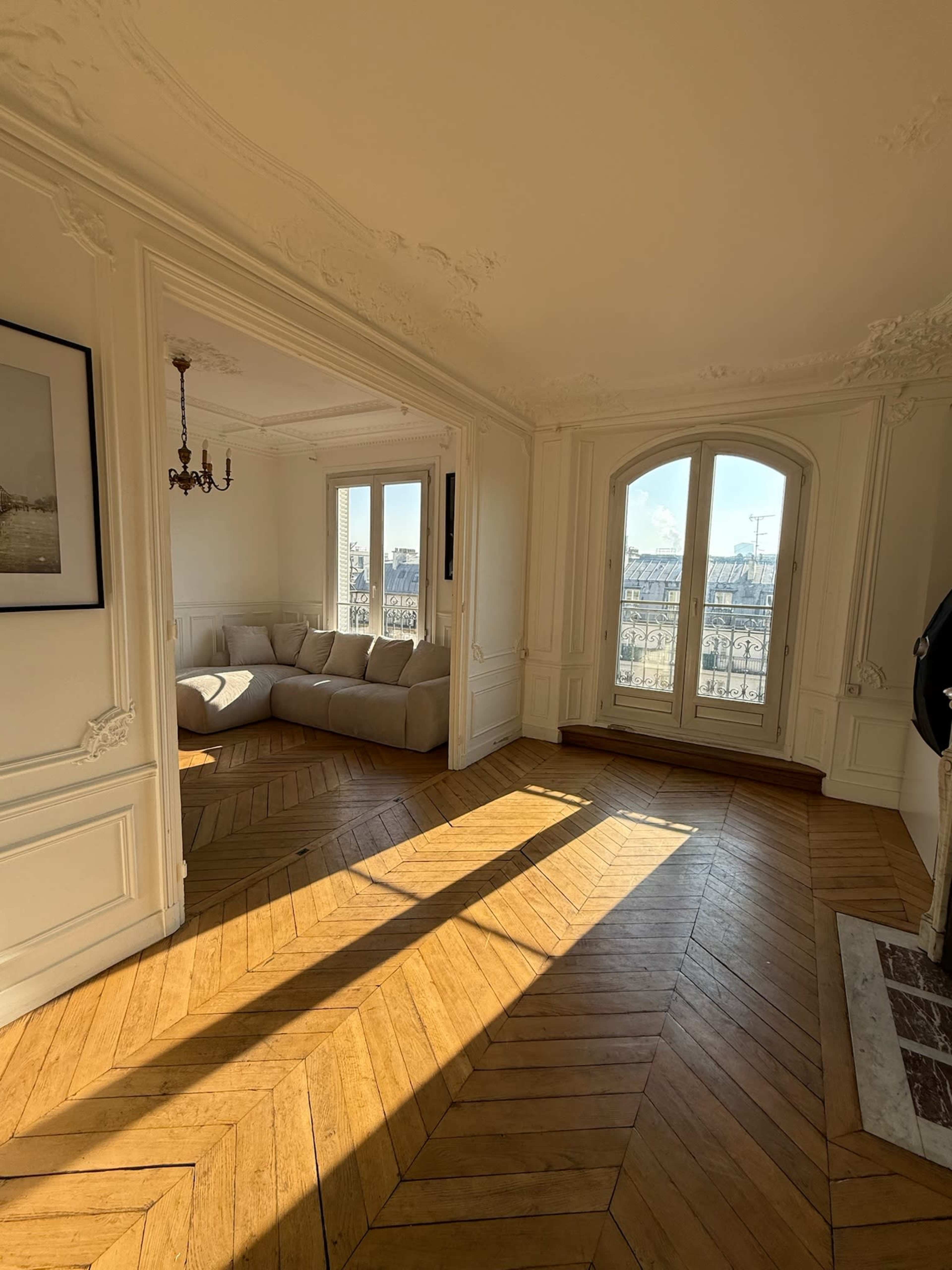 A well-lit room featuring a beige sofa, intricate molding, and a large window with a view of the city.