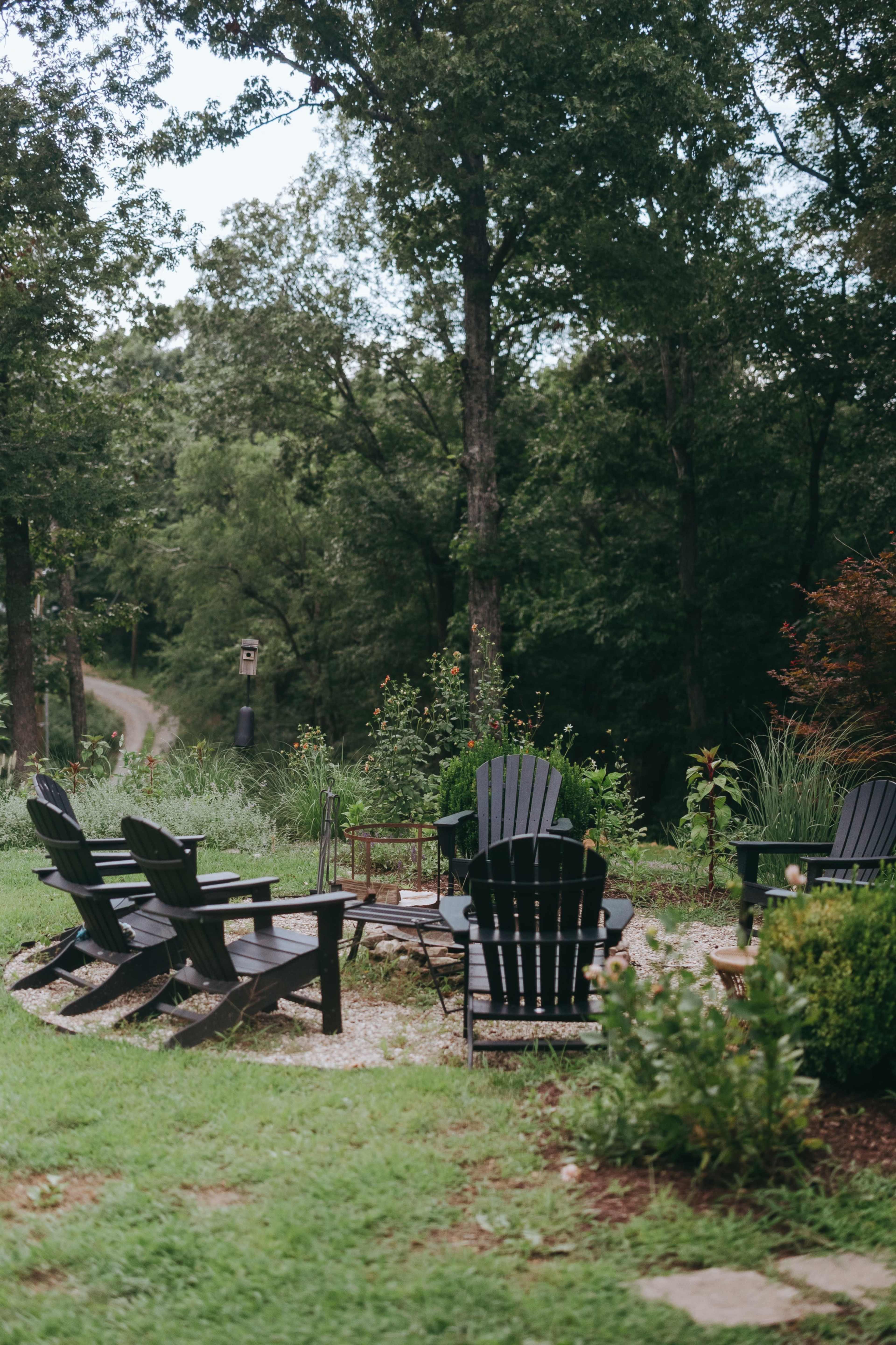 A circle of black Adirondack chairs is arranged around a small stone fire pit, with a lush landscape of trees and greenery in the background.