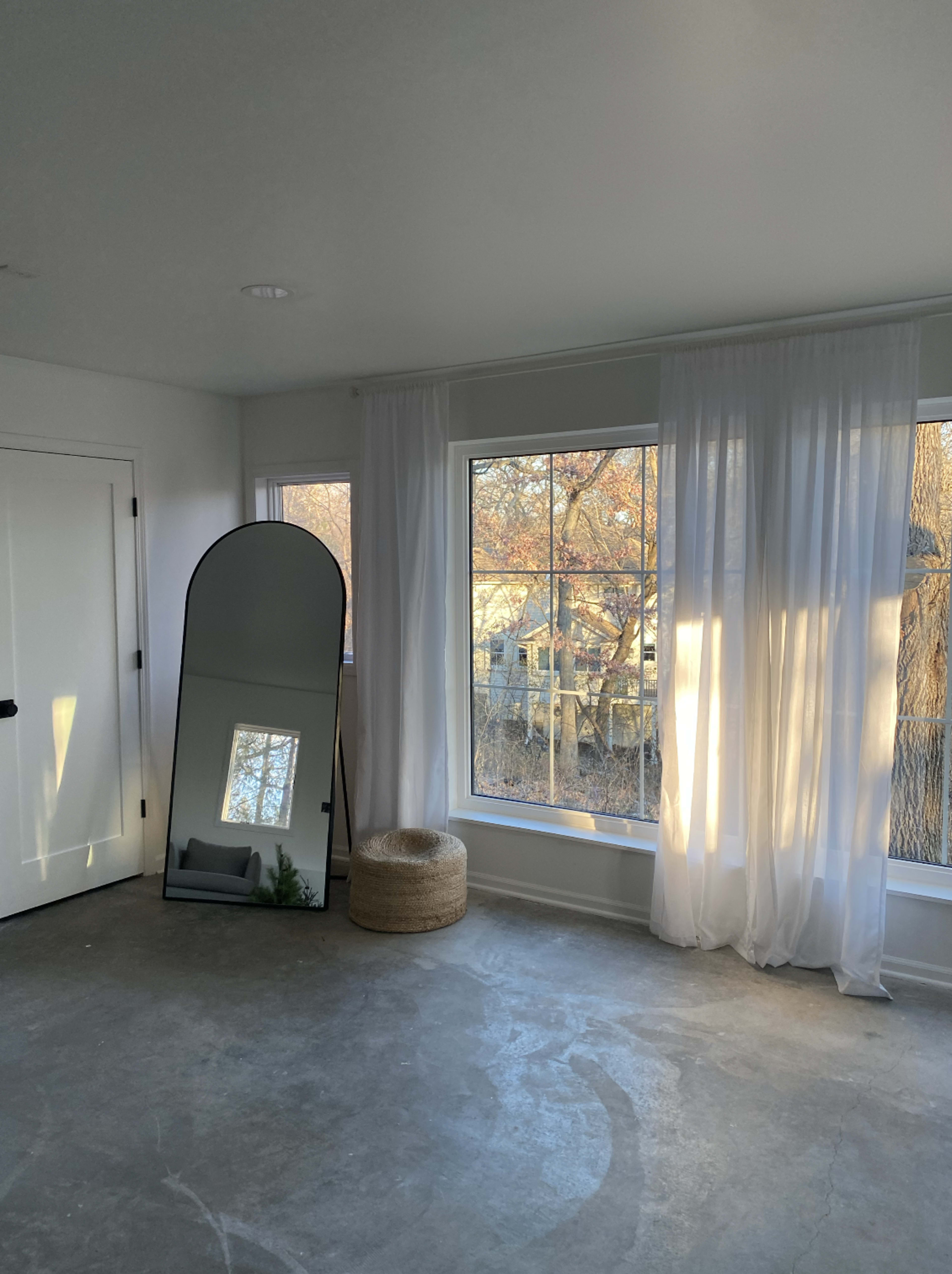 A minimalist room features a tall mirror, large windows with sheer curtains, and a round woven basket.