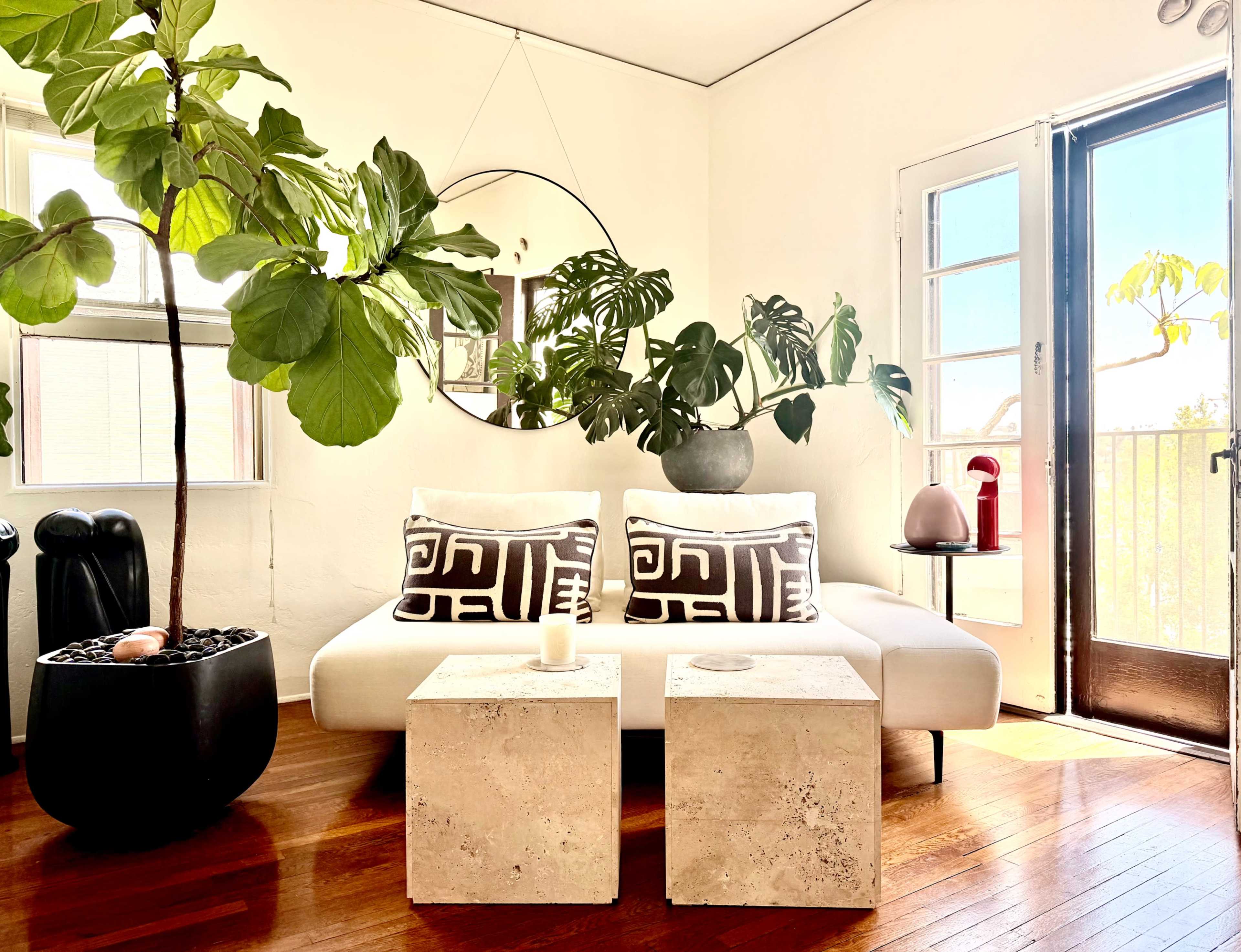 The image shows a cozy living room with a white sofa, two stone coffee tables, large indoor plants, and a round mirror reflecting the light from a nearby window.