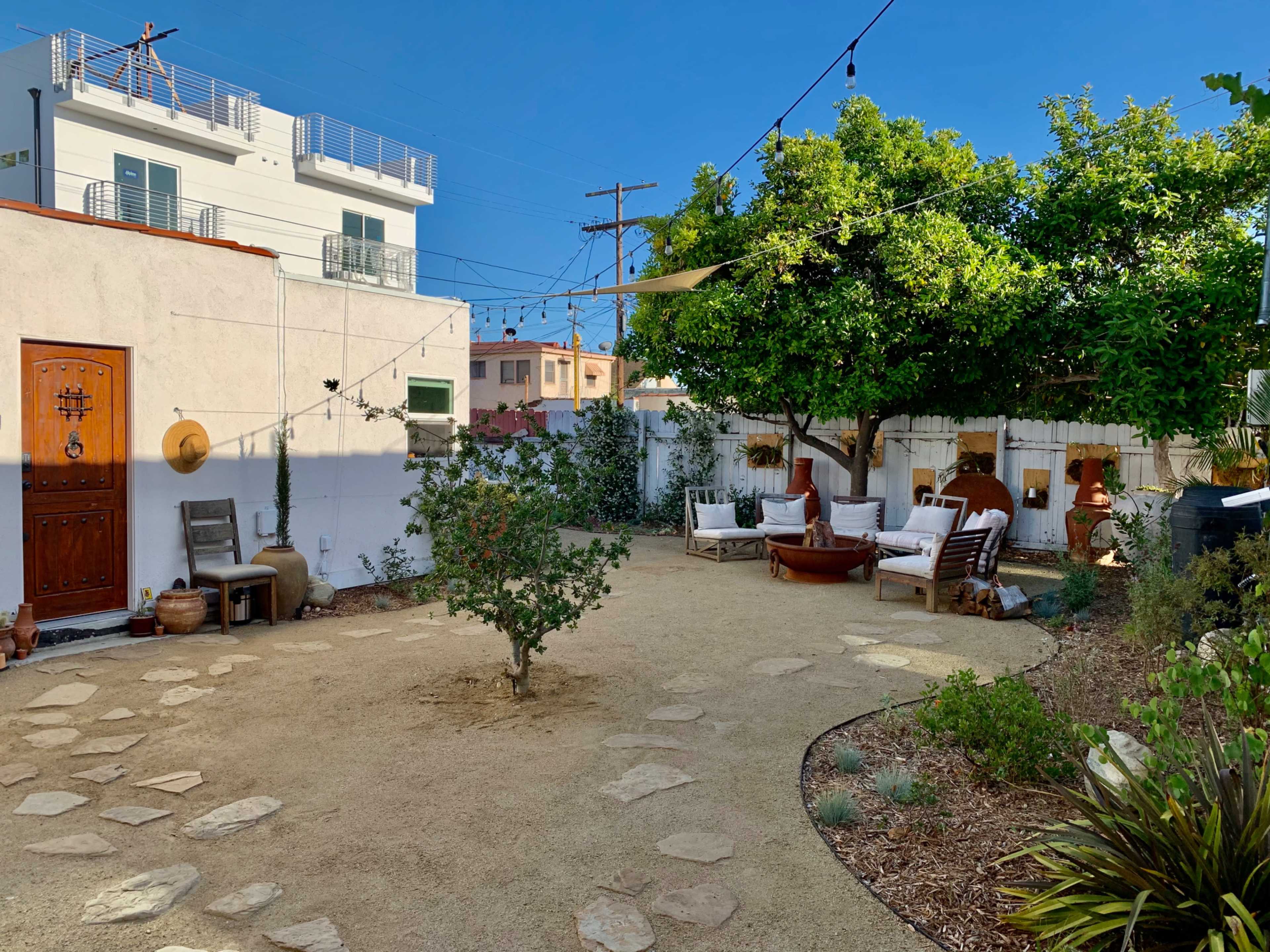 A small outdoor courtyard with gravel ground, stone pathways, seating areas made of wood, and a citrus tree surrounded by various plants.