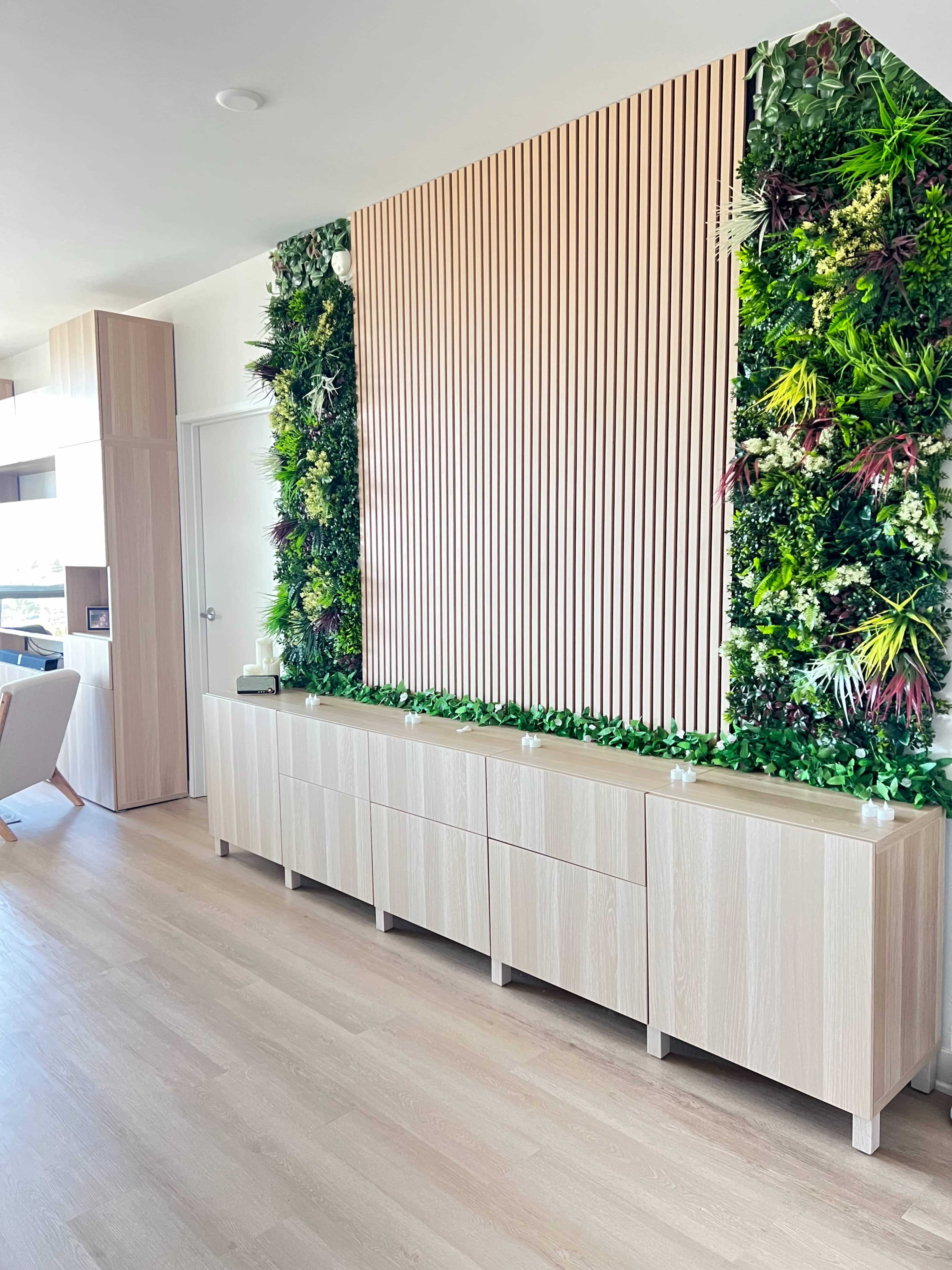The image shows a modern living space featuring a light wooden cabinet against a vibrant vertical garden wall adorned with various green plants.
