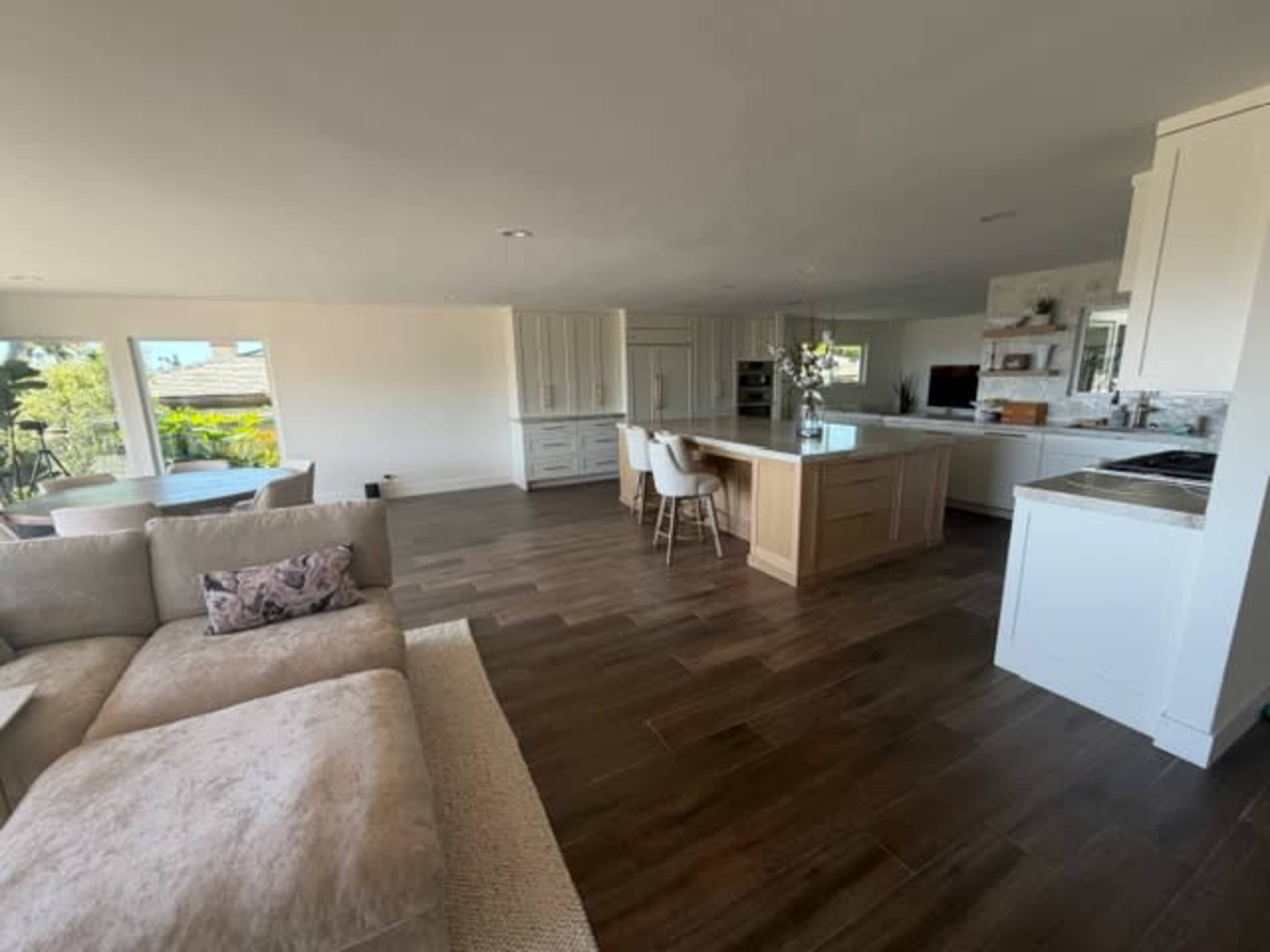 The image shows a modern kitchen and living area with large windows, wooden flooring, and a large island with bar stools.