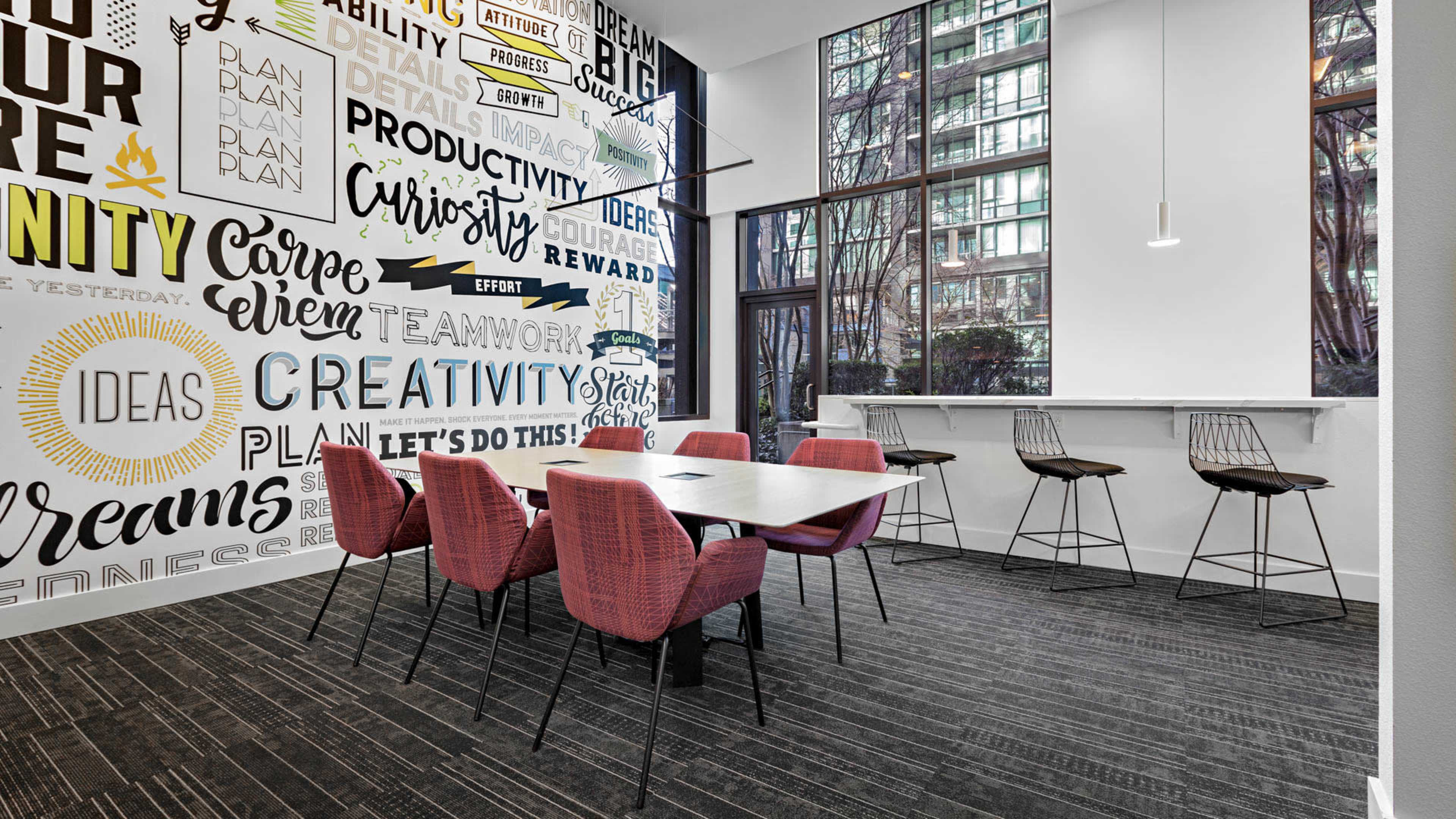 A modern workspace features a table with six red chairs, a bar with high stools, and a wall adorned with motivational words and phrases.