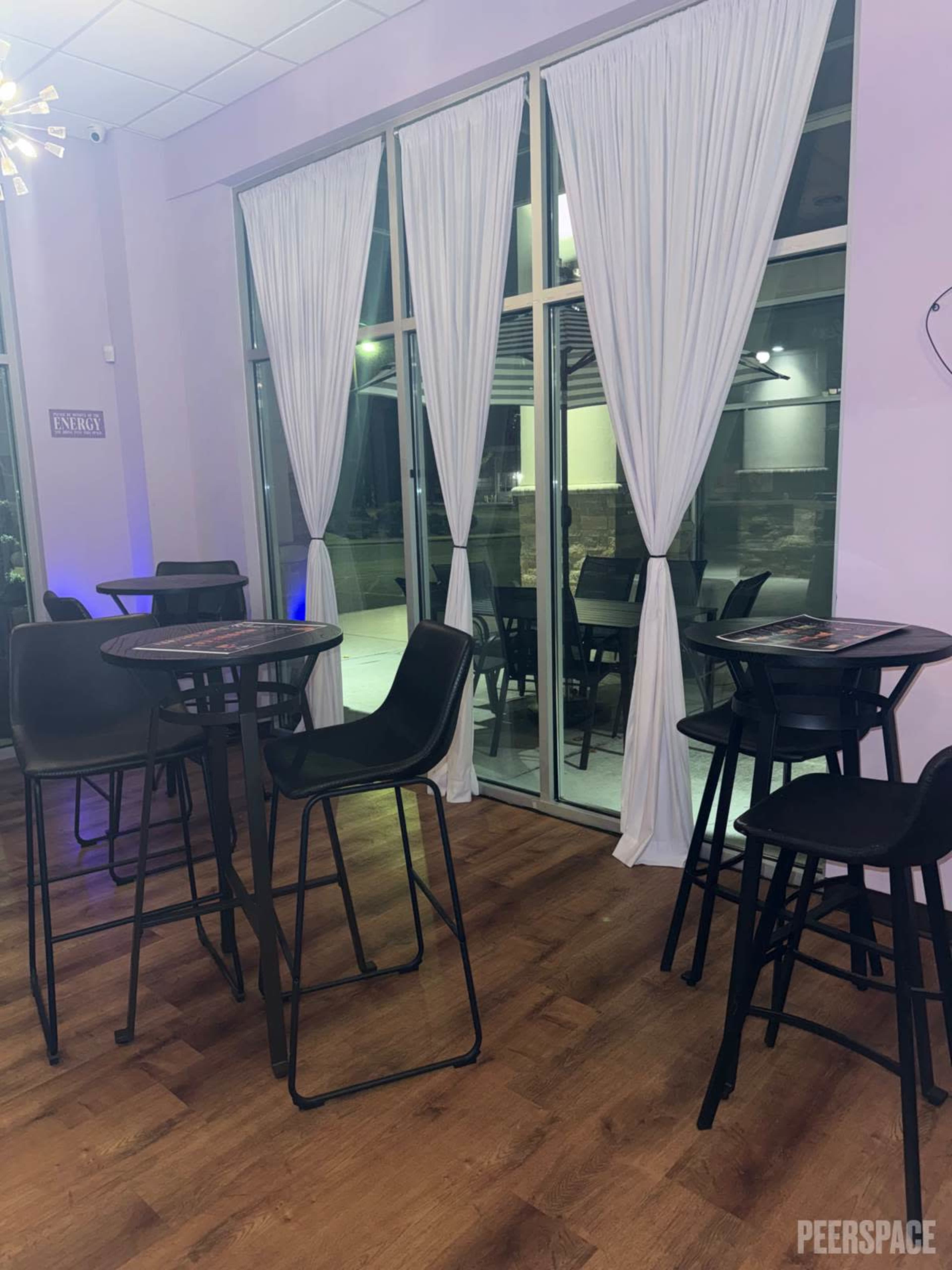 The image shows a modern indoor space featuring high tables and black chairs, with large windows covered by white sheer curtains.