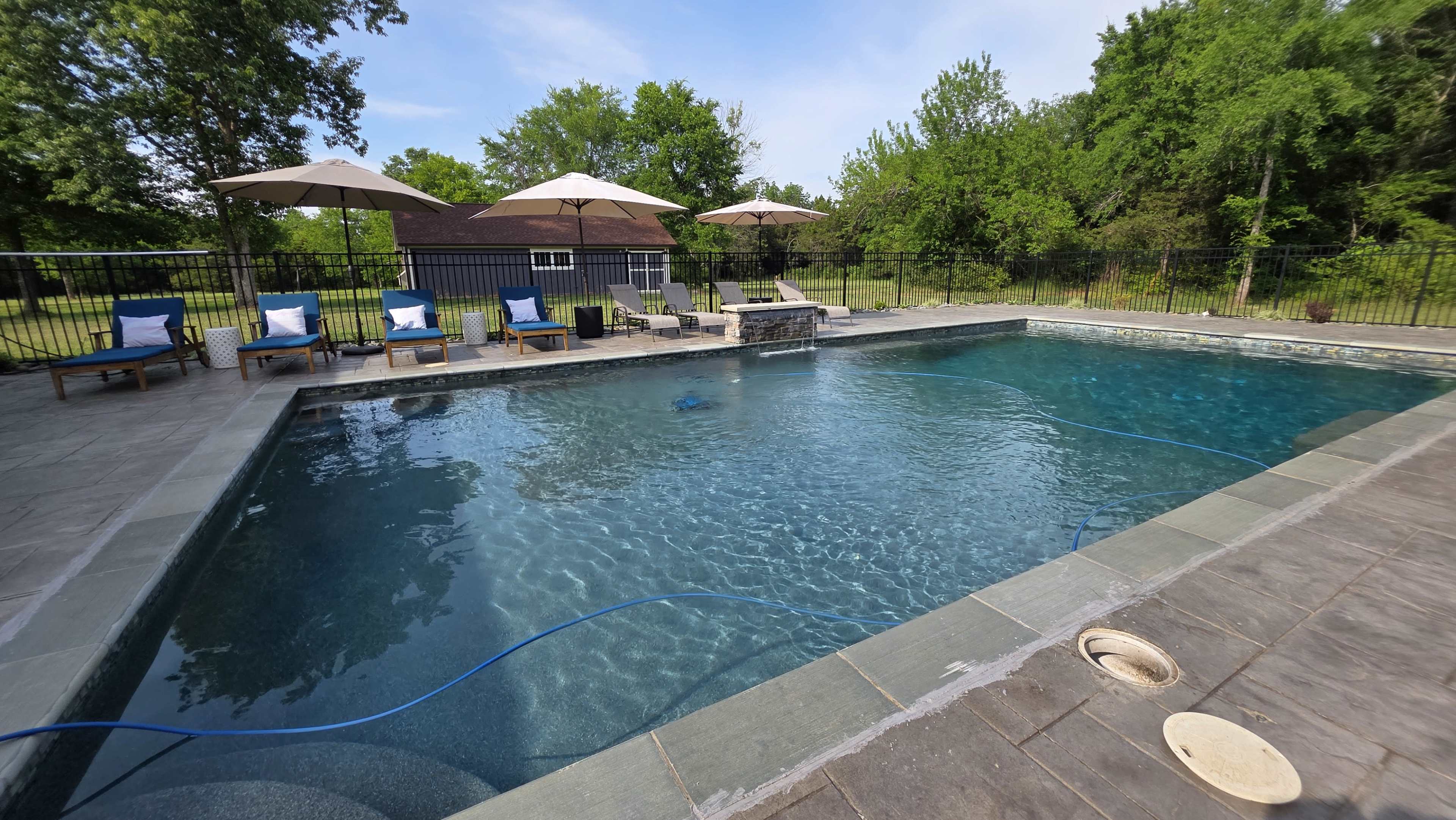 The image depicts a swimming pool surrounded by lounge chairs and umbrellas, with green trees and a house in the background.