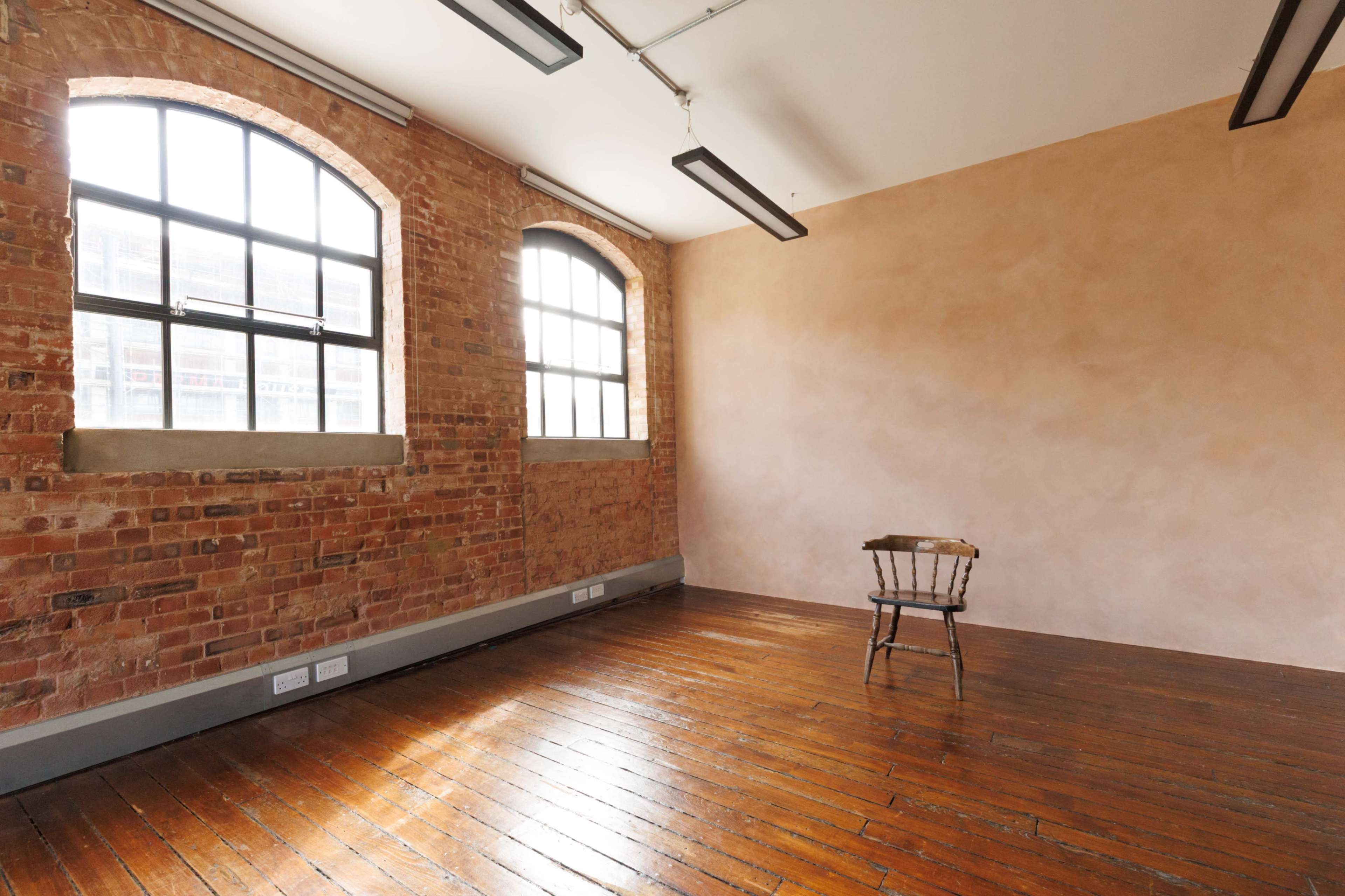 The image shows an empty room with exposed brick walls, wooden flooring, and a single chair placed in the center under large windows.