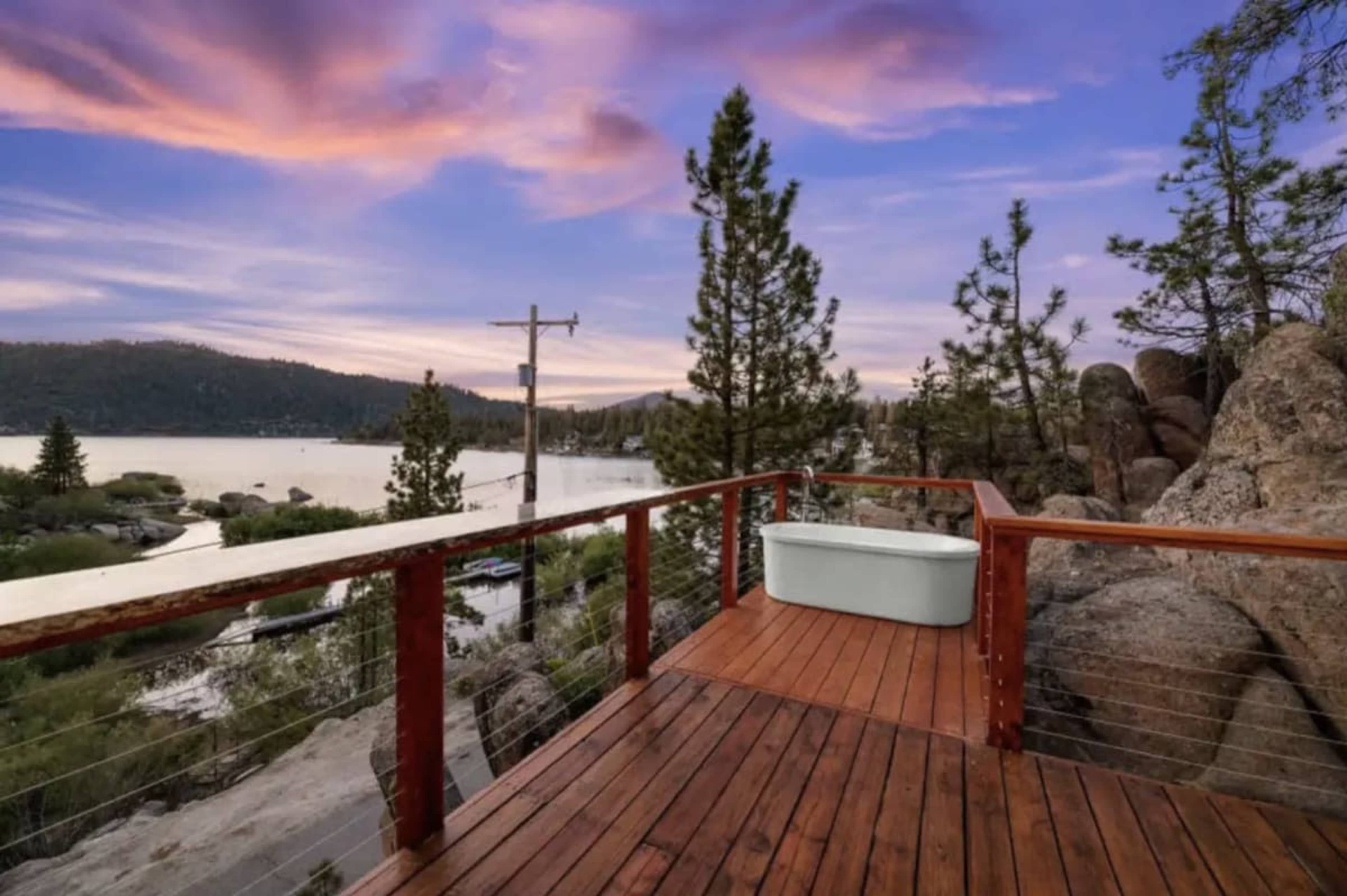 A wooden deck overlooks a tranquil lake surrounded by trees and rocks under a colorful sunset sky.