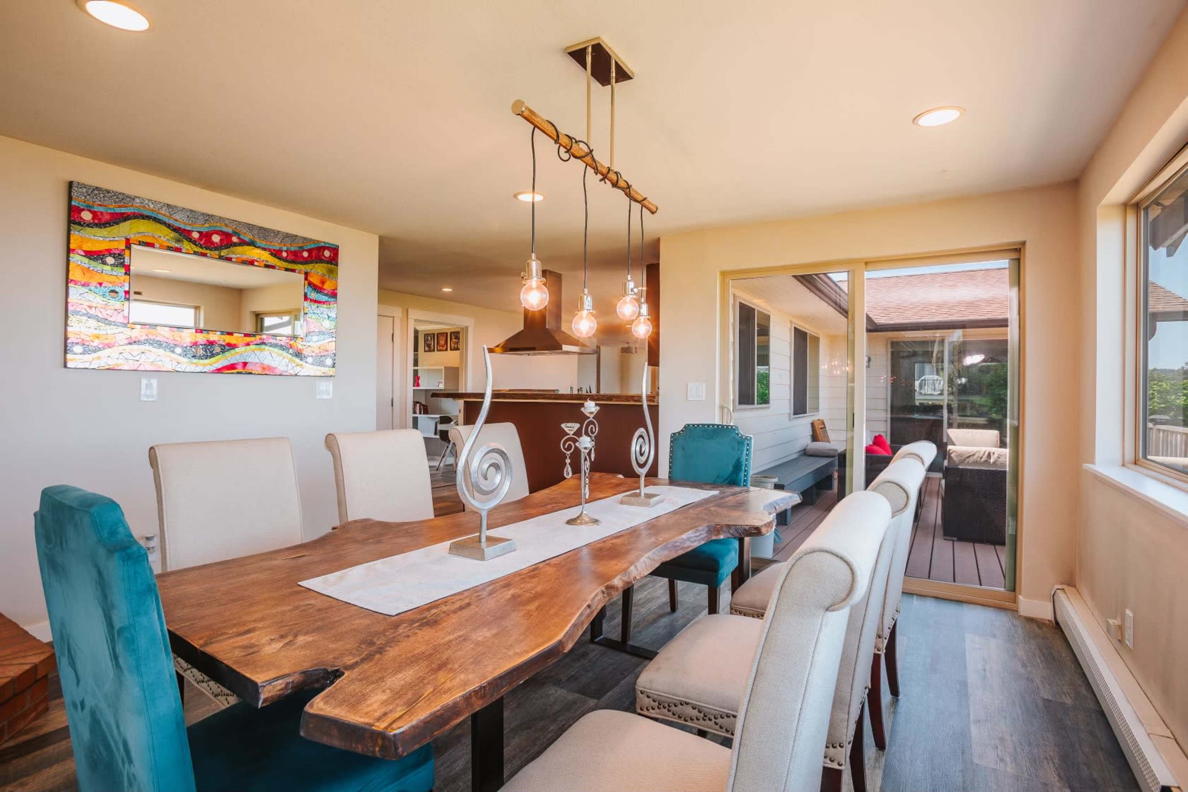 A spacious dining area features a large wooden table surrounded by upholstered chairs, with modern light fixtures and a colorful wall mirror.