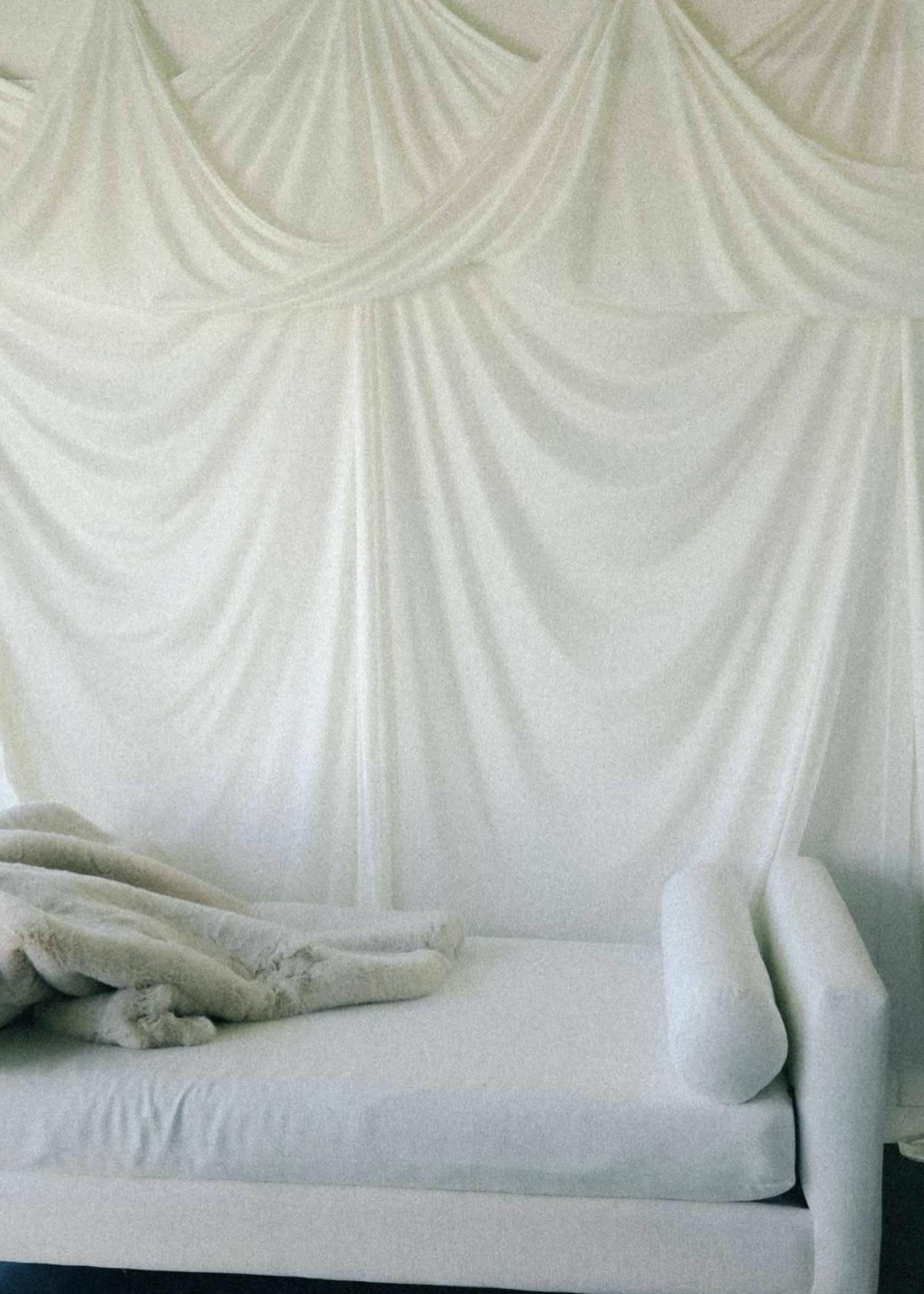 The image shows a minimalistic white couch draped with a soft blanket against a backdrop of flowing white fabric.