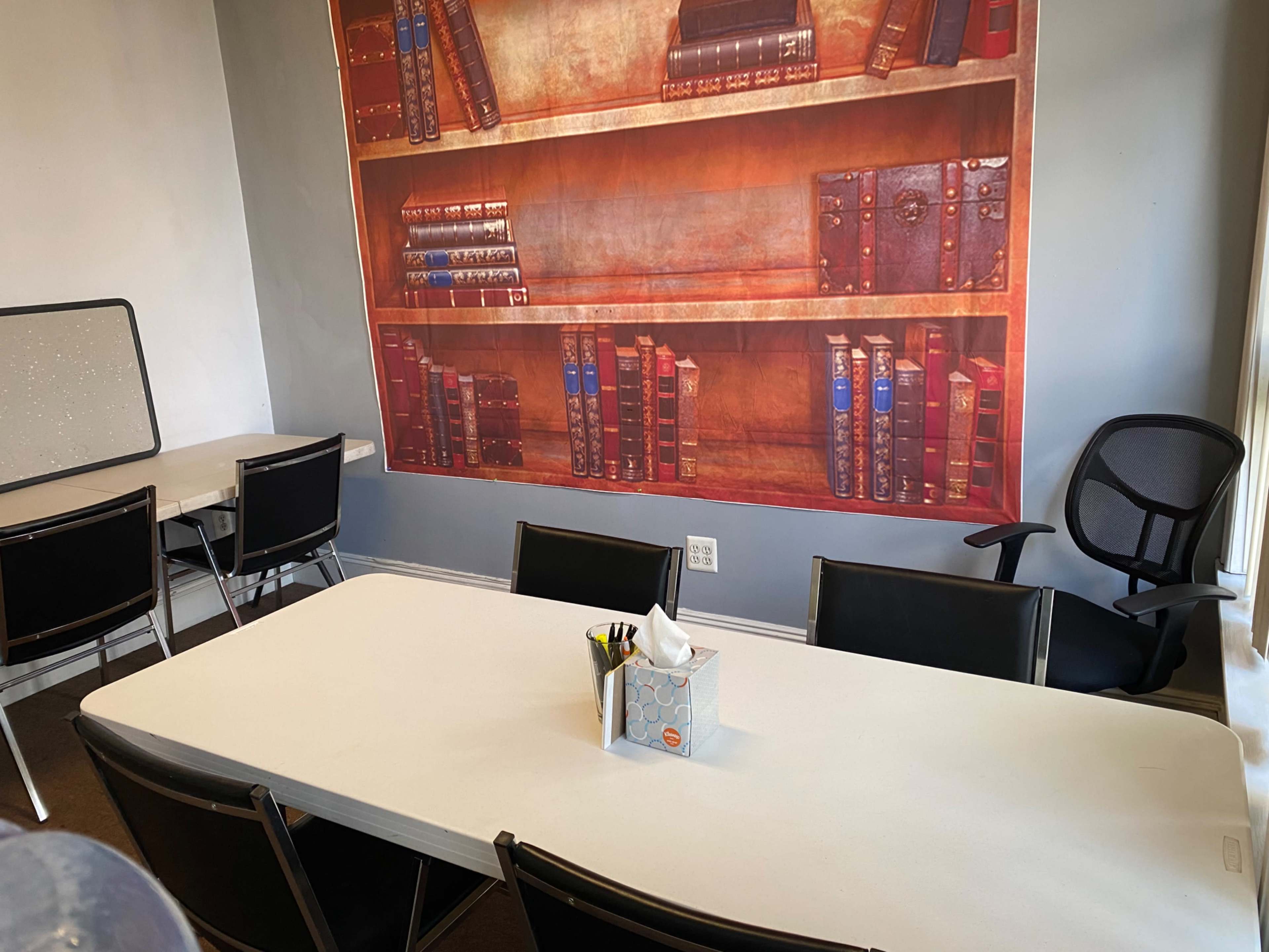 A meeting room features a long table with black chairs, a tissue box, and a mural of bookshelves on the wall.