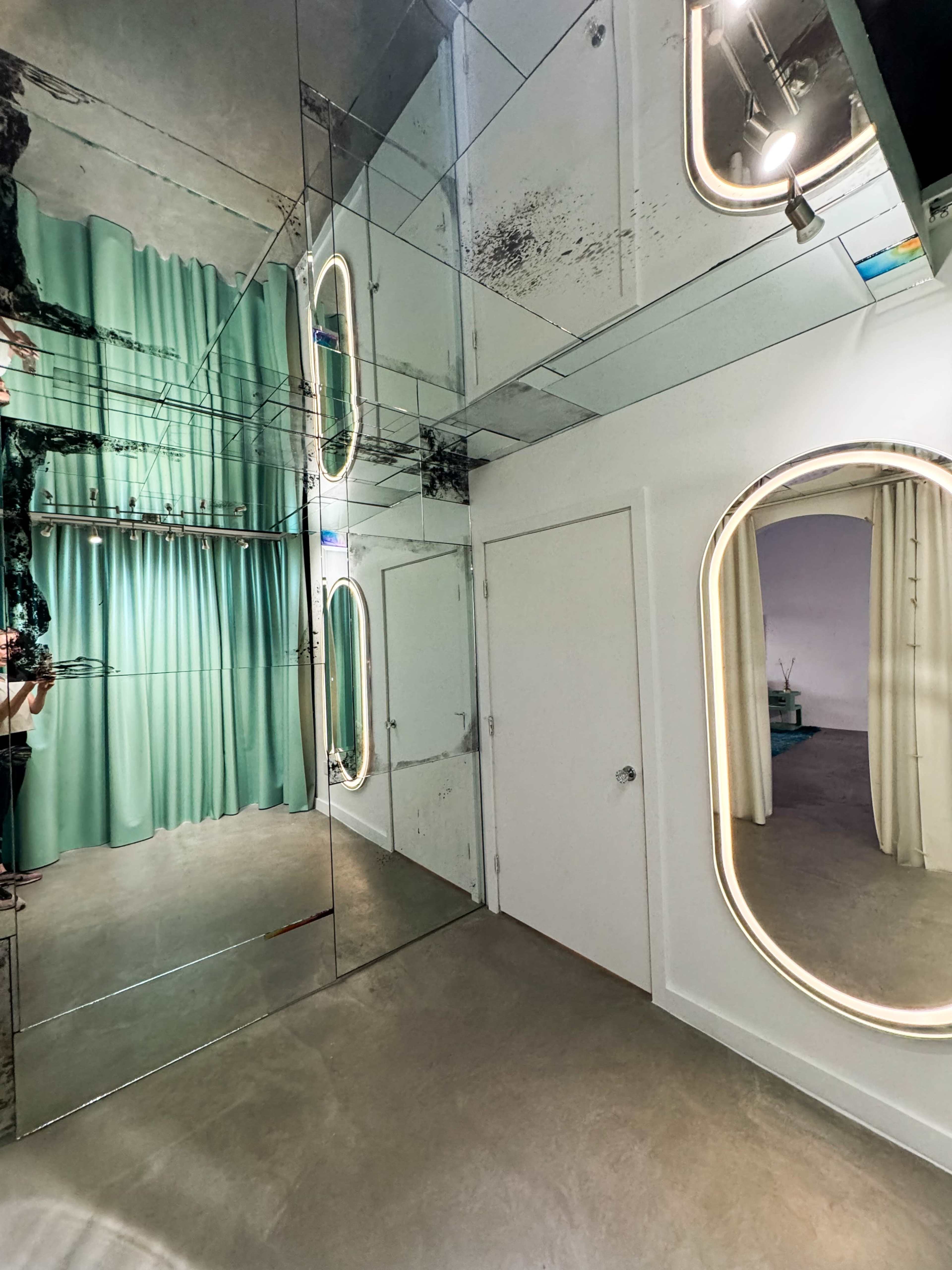 The image shows a modern interior space featuring mirrored walls, green drapery, and a doorway with a rounded mirror.