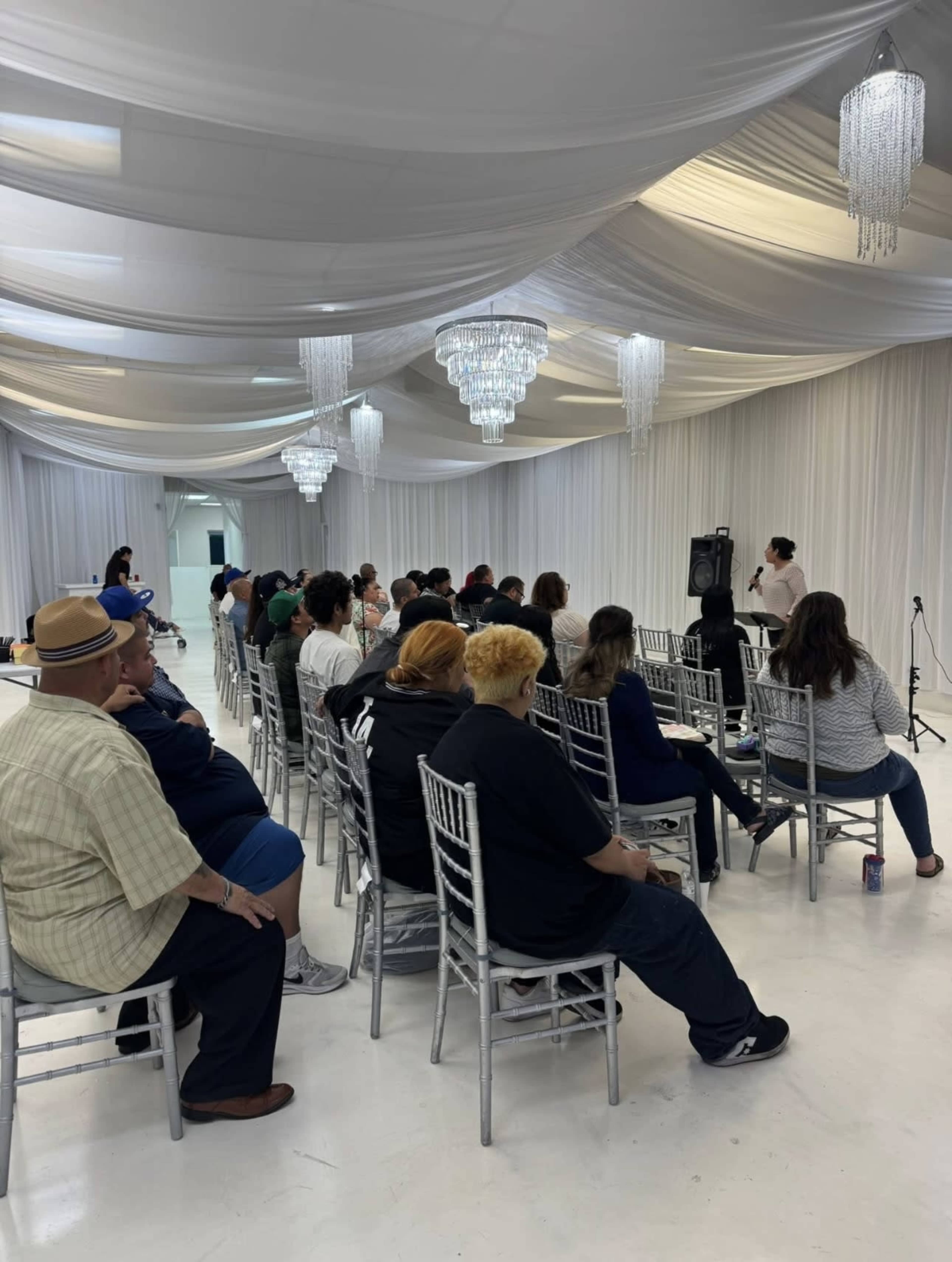 A speaker addresses an audience seated in chairs under a decorative ceiling with chandeliers in a well-lit event space.