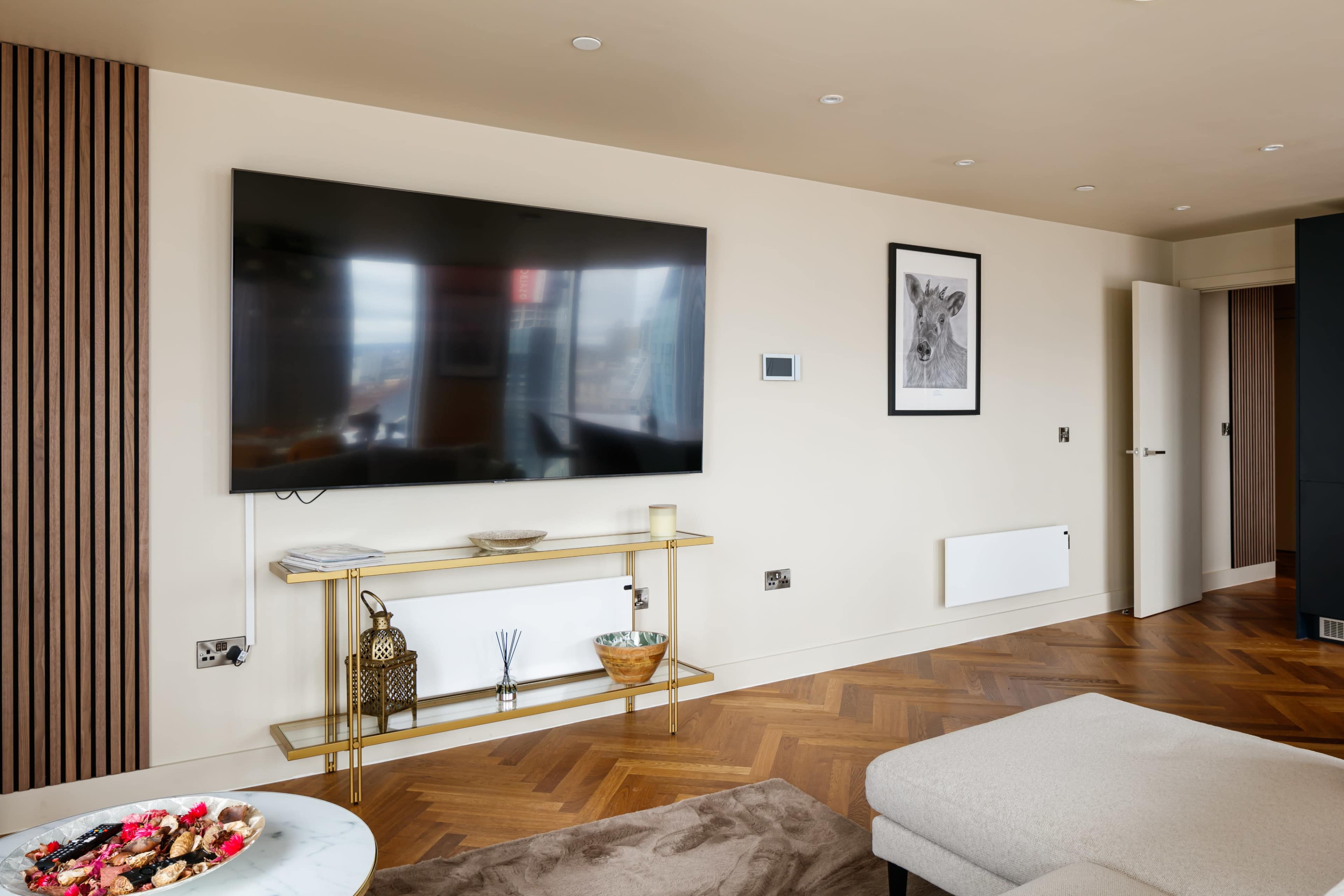The image shows a modern living room featuring a large flat-screen TV mounted on the wall above a golden console table, with a sofa and wooden flooring.
