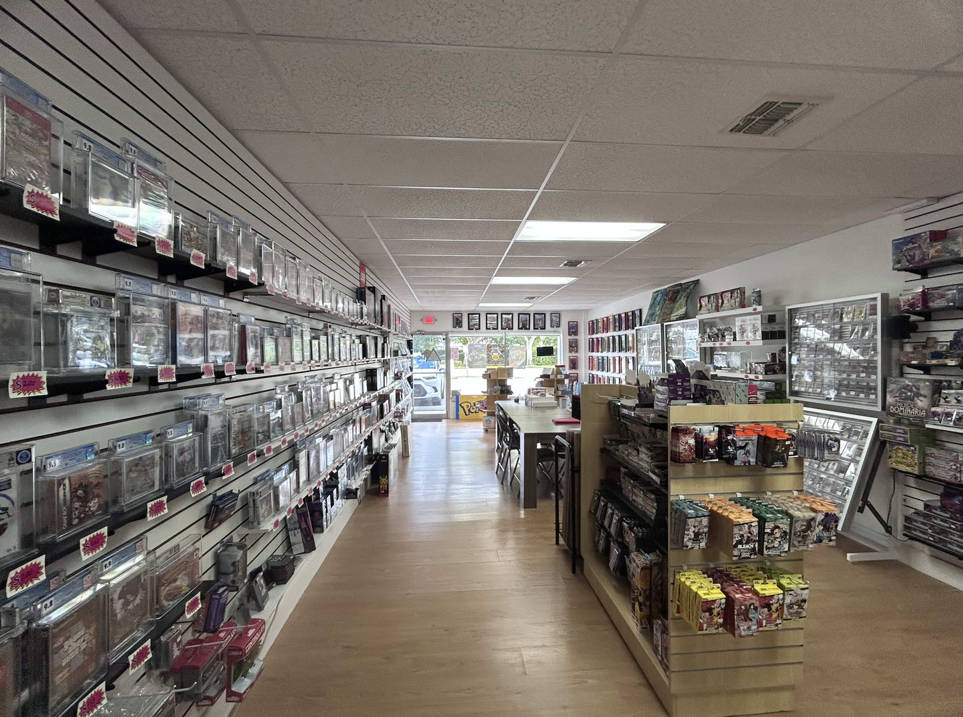 The image shows the interior of a store with shelves lined with packaged collectibles and trading cards, a central display table, and bright lighting.
