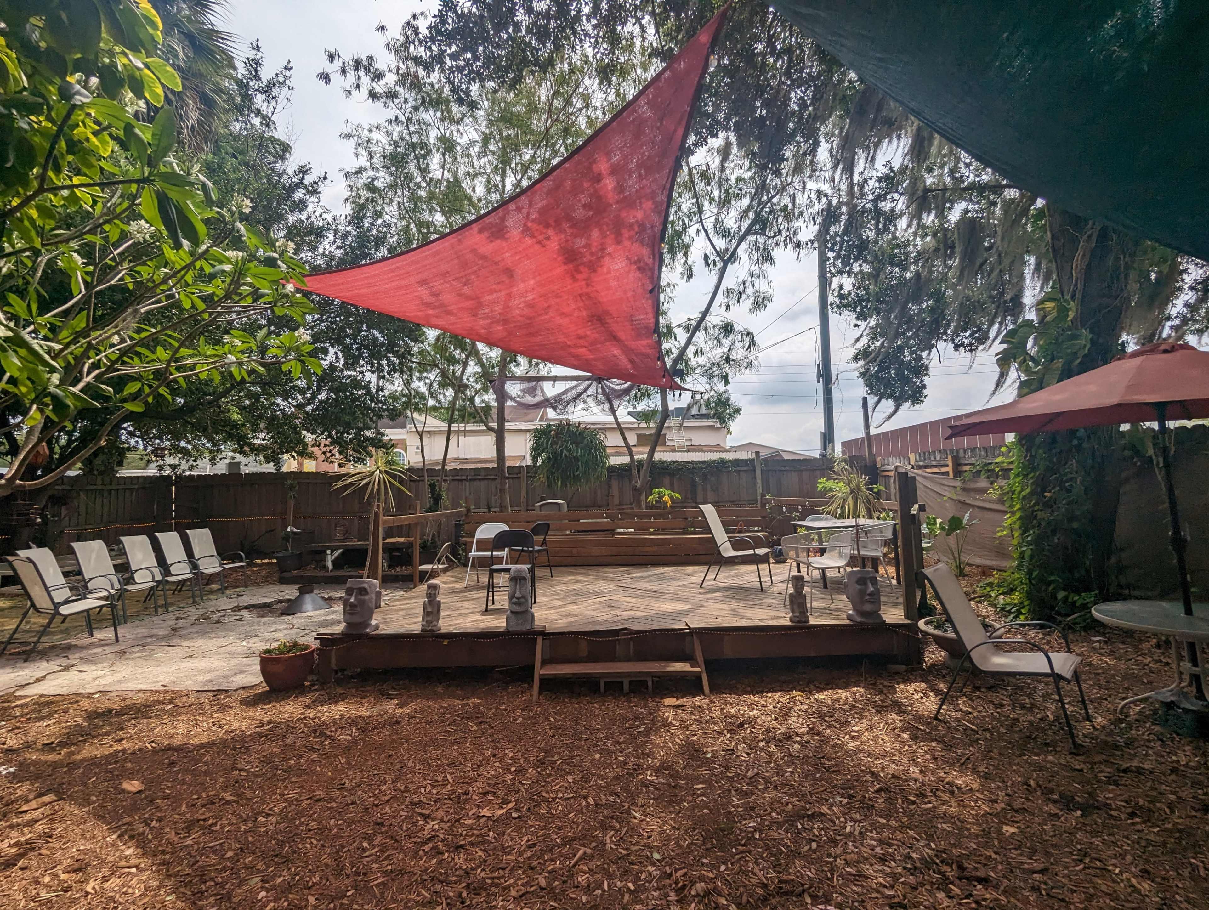 The image shows a backyard space with wooden decking, several chairs arranged around, and red shading fabric overhead amidst greenery and decorative elements.