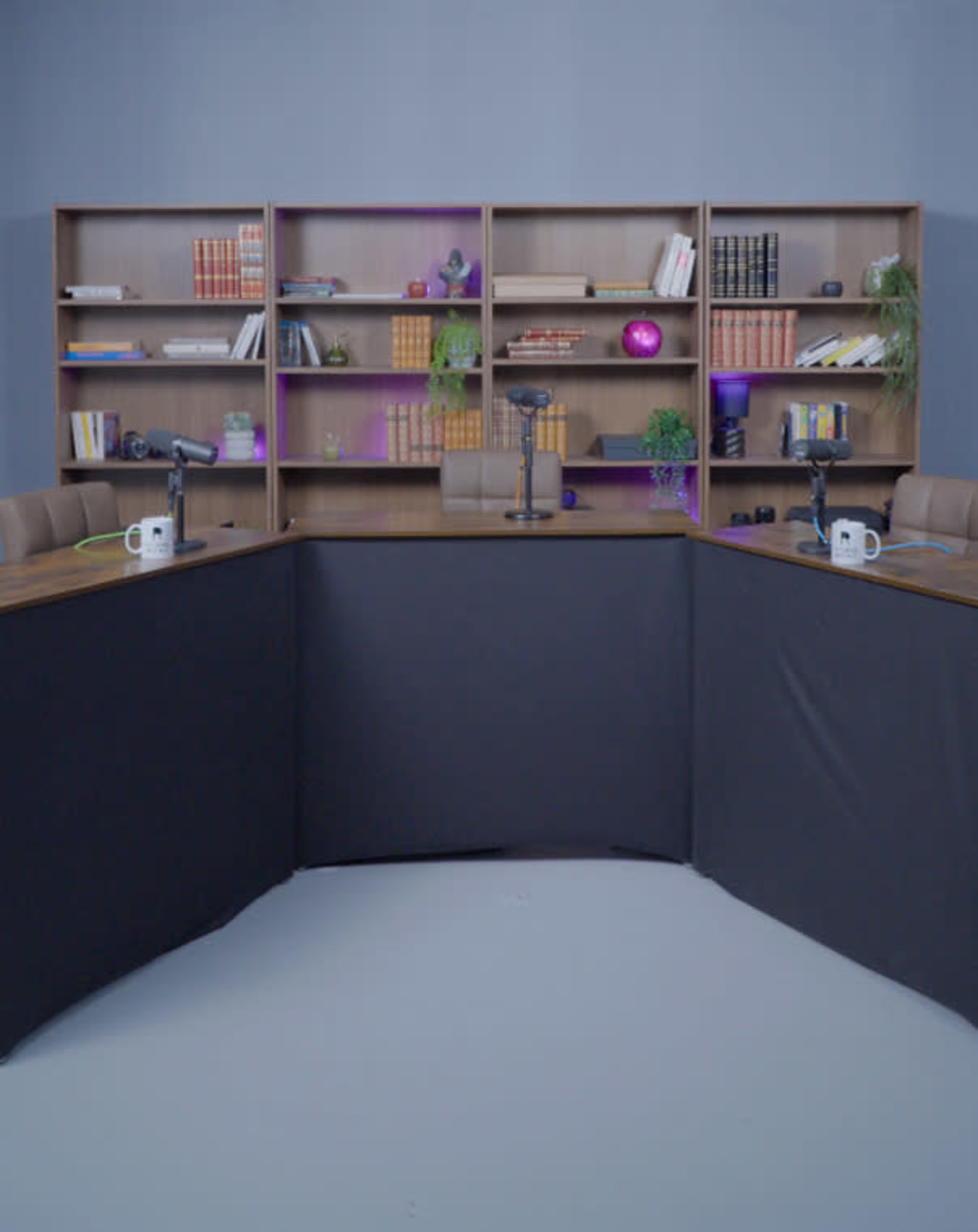 The image shows a podcast recording studio with two microphones, a round table covered with a black cloth, and a bookshelf filled with various books and decorative items in the background.