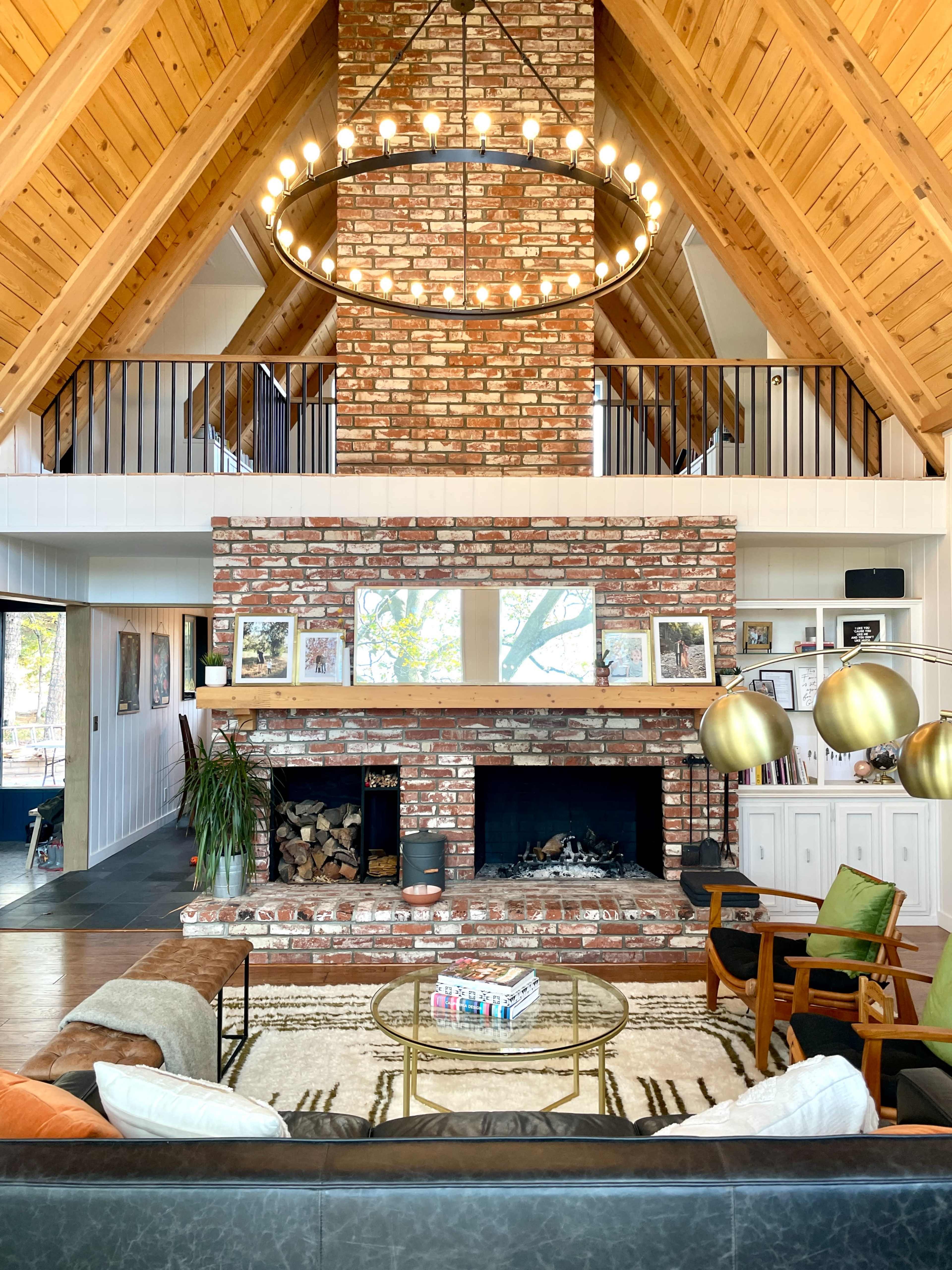 The image shows a spacious living room with a high wooden ceiling, a large brick fireplace, and a circular chandelier hanging above a coffee table situated on a patterned rug.