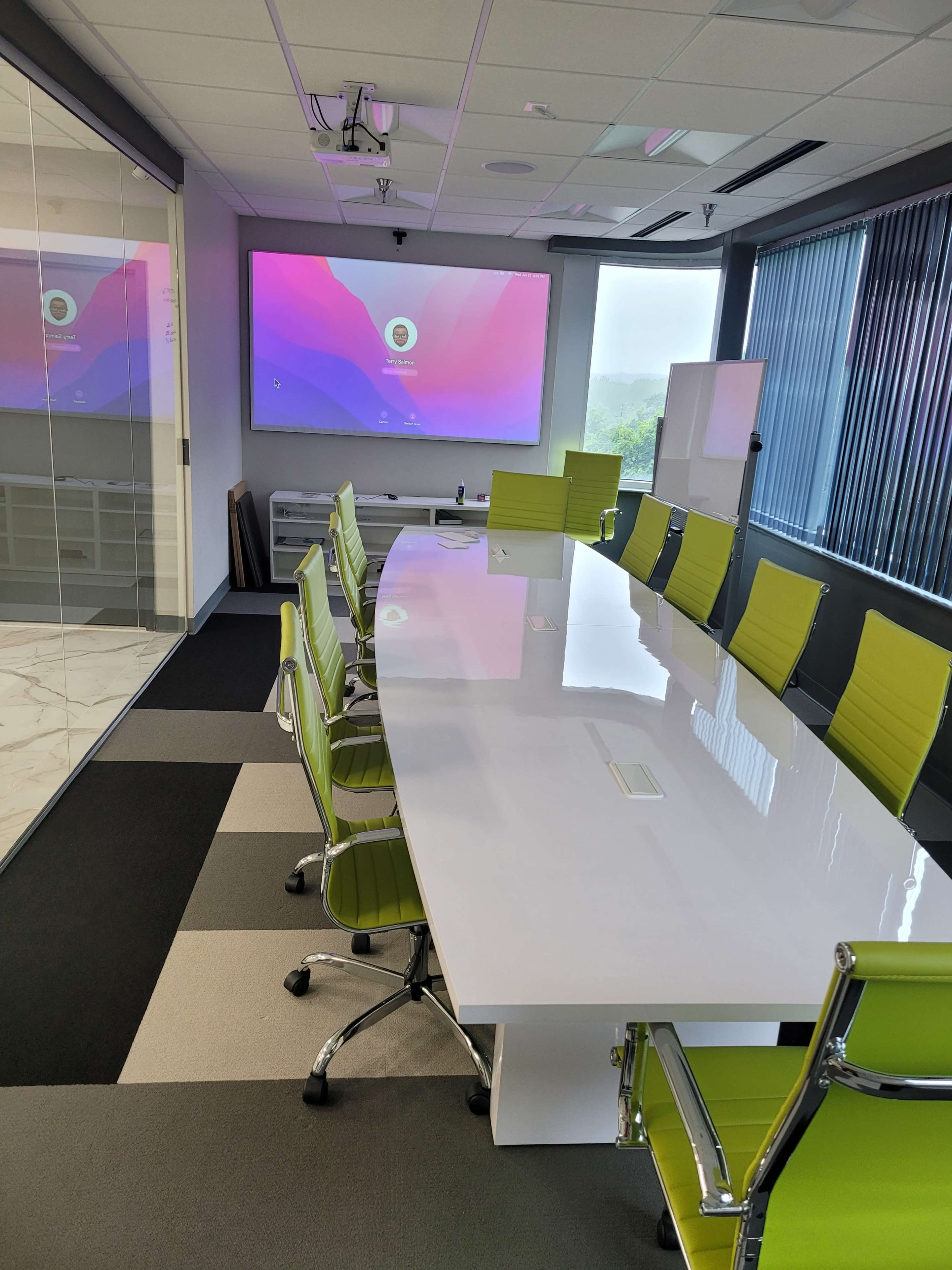 A modern conference room features a long white table surrounded by green chairs, with a large screen displaying a colorful presentation in the background.