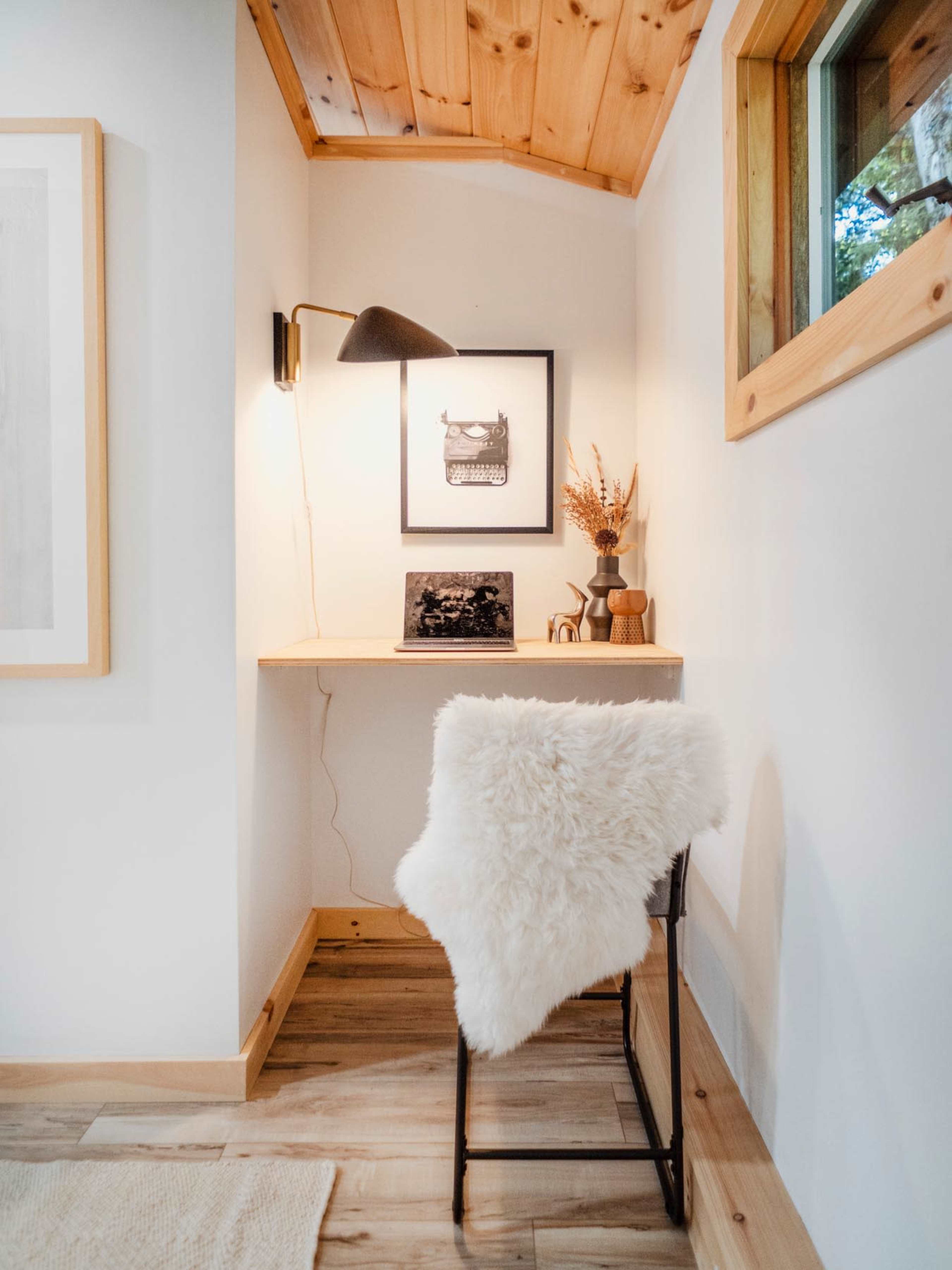 A small workspace features a desk with an illuminated lamp, an adorned wall with framed art, and a chair draped with a fluffy throw.