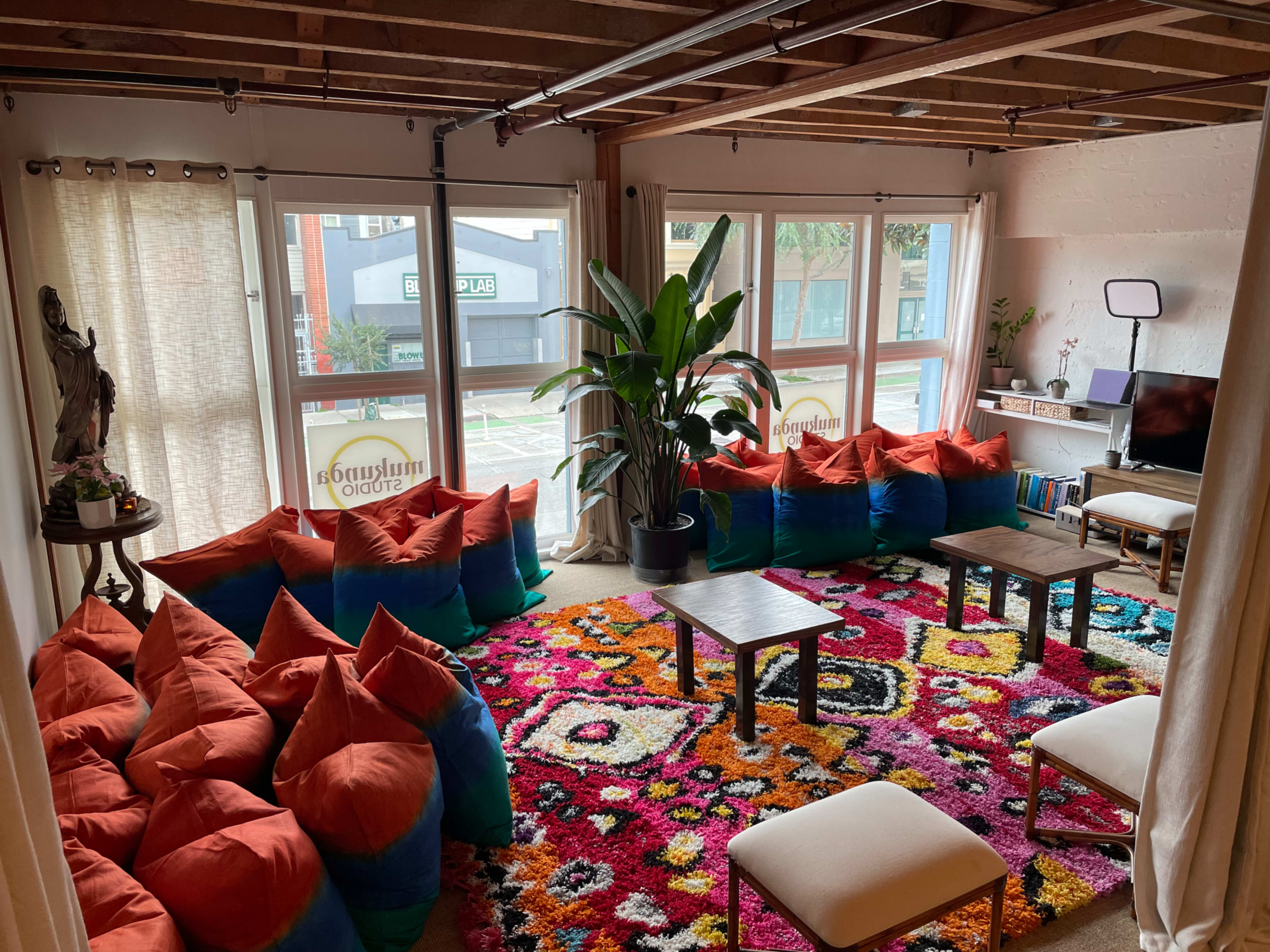The image shows a cozy lounge area with a colorful area rug, bean bag seating, a small coffee table, and large windows allowing natural light.
