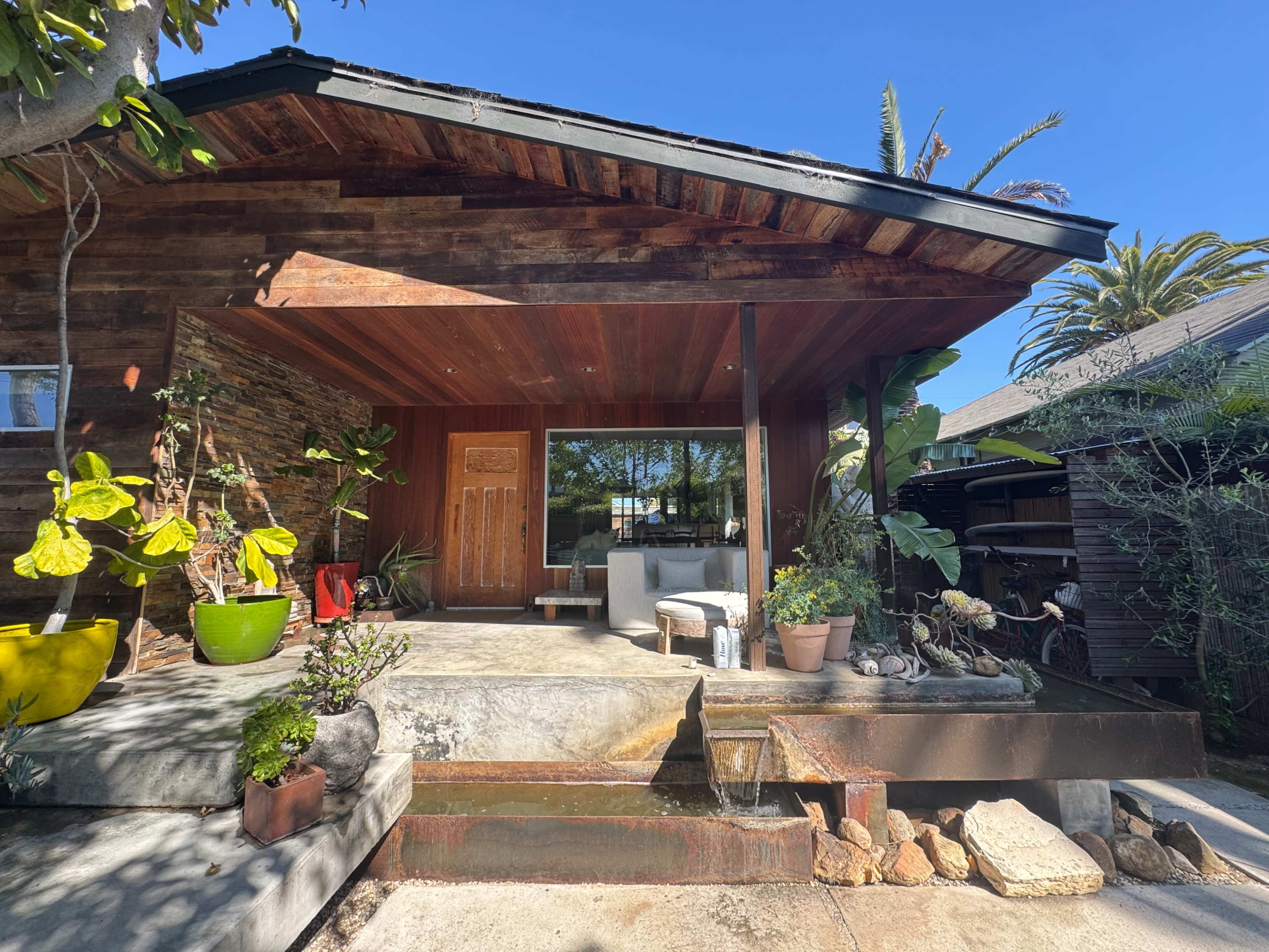 The image shows a wooden-fronted house with a small seating area and various potted plants lining a stone pathway.