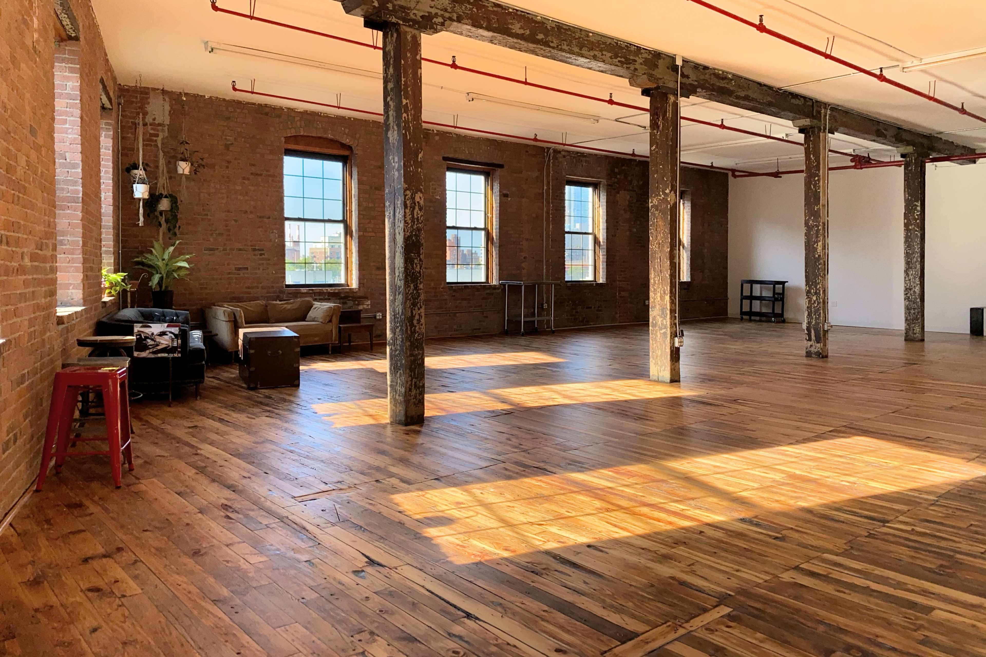The image shows a spacious, well-lit room with brick walls, wooden floors, and exposed beams, featuring a couch, a coffee table, and large windows.