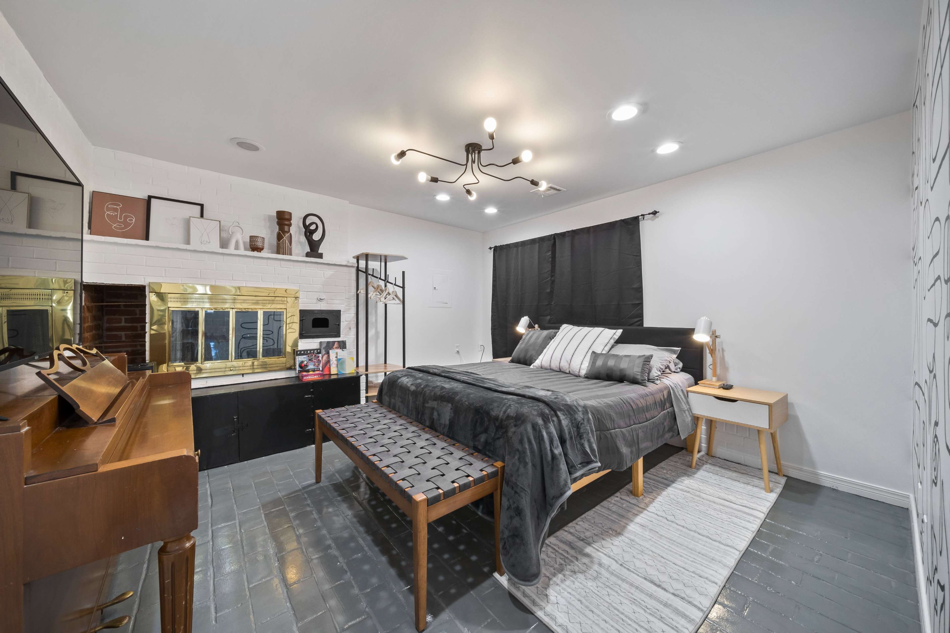 The image shows a modern bedroom featuring a bed with a gray throw blanket, a wooden bench at the foot, and a fireplace surrounded by black cabinetry.