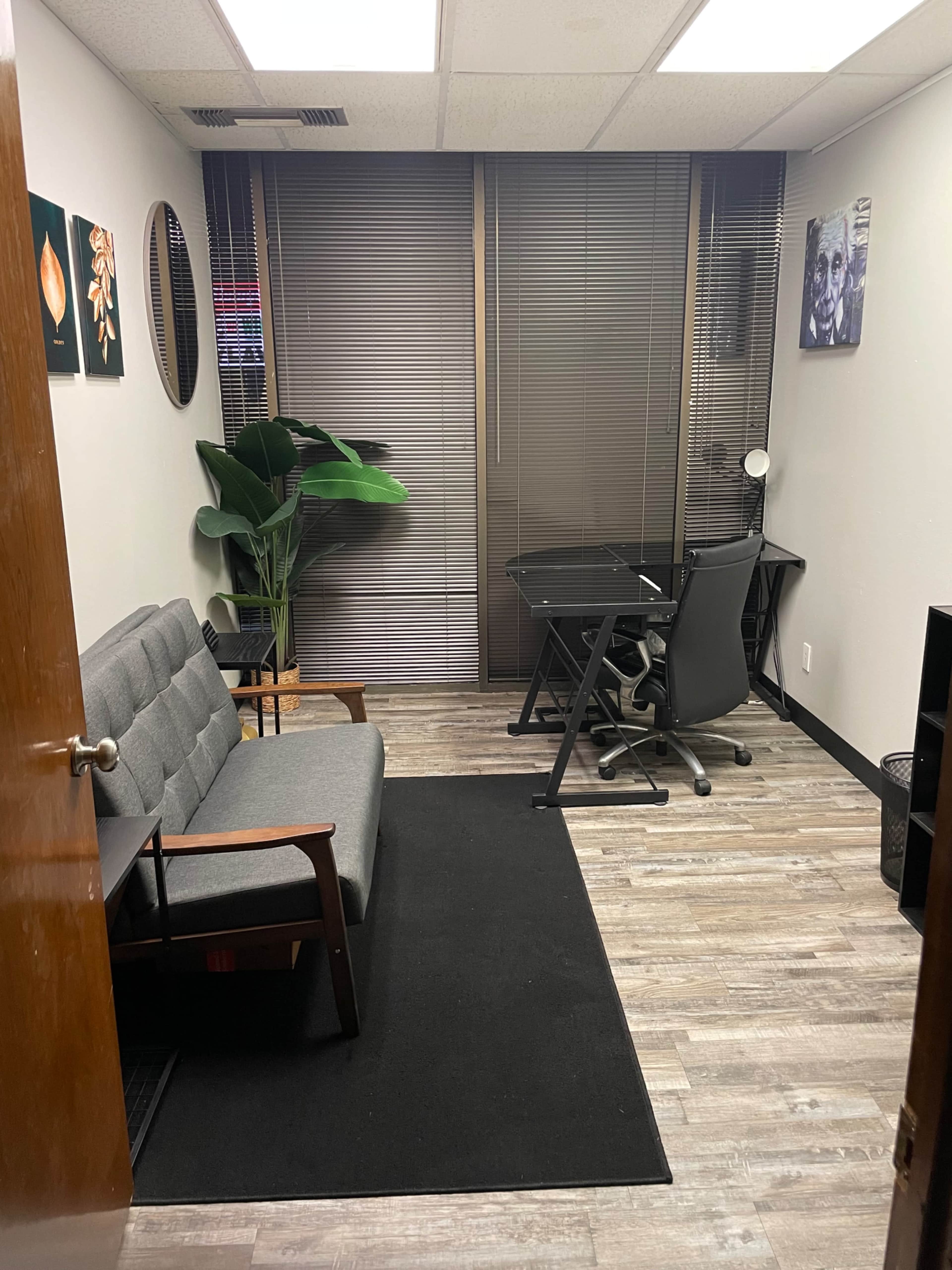 The image shows a small office space with a gray couch, a desk with a chair, and a large plant near a window with blinds.