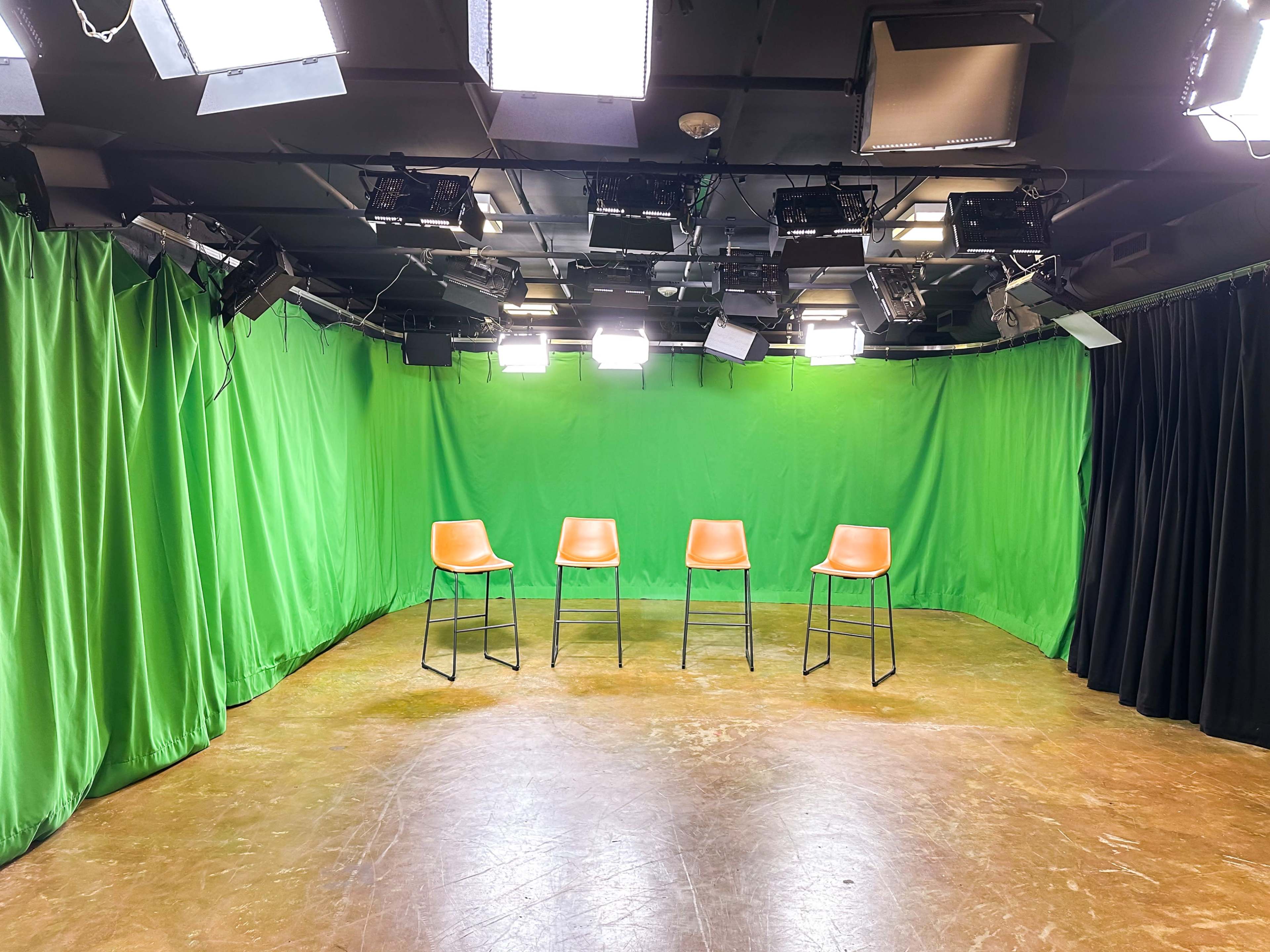 A television studio setup with four empty orange bar stools on a bare floor surrounded by green and black curtains and bright overhead lights.