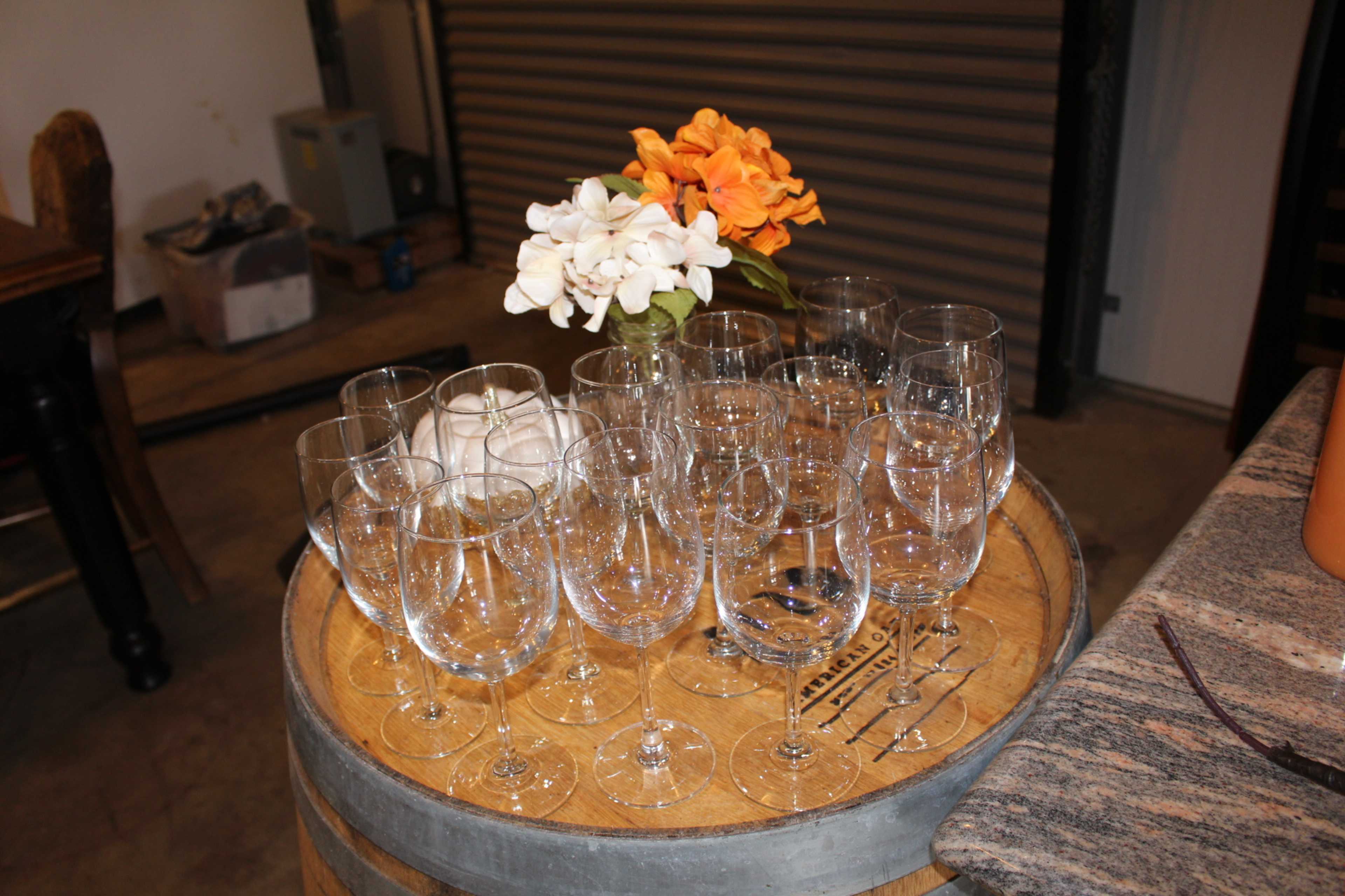 A collection of various glassware is arranged on a wooden barrel, accompanied by a small vase of flowers.